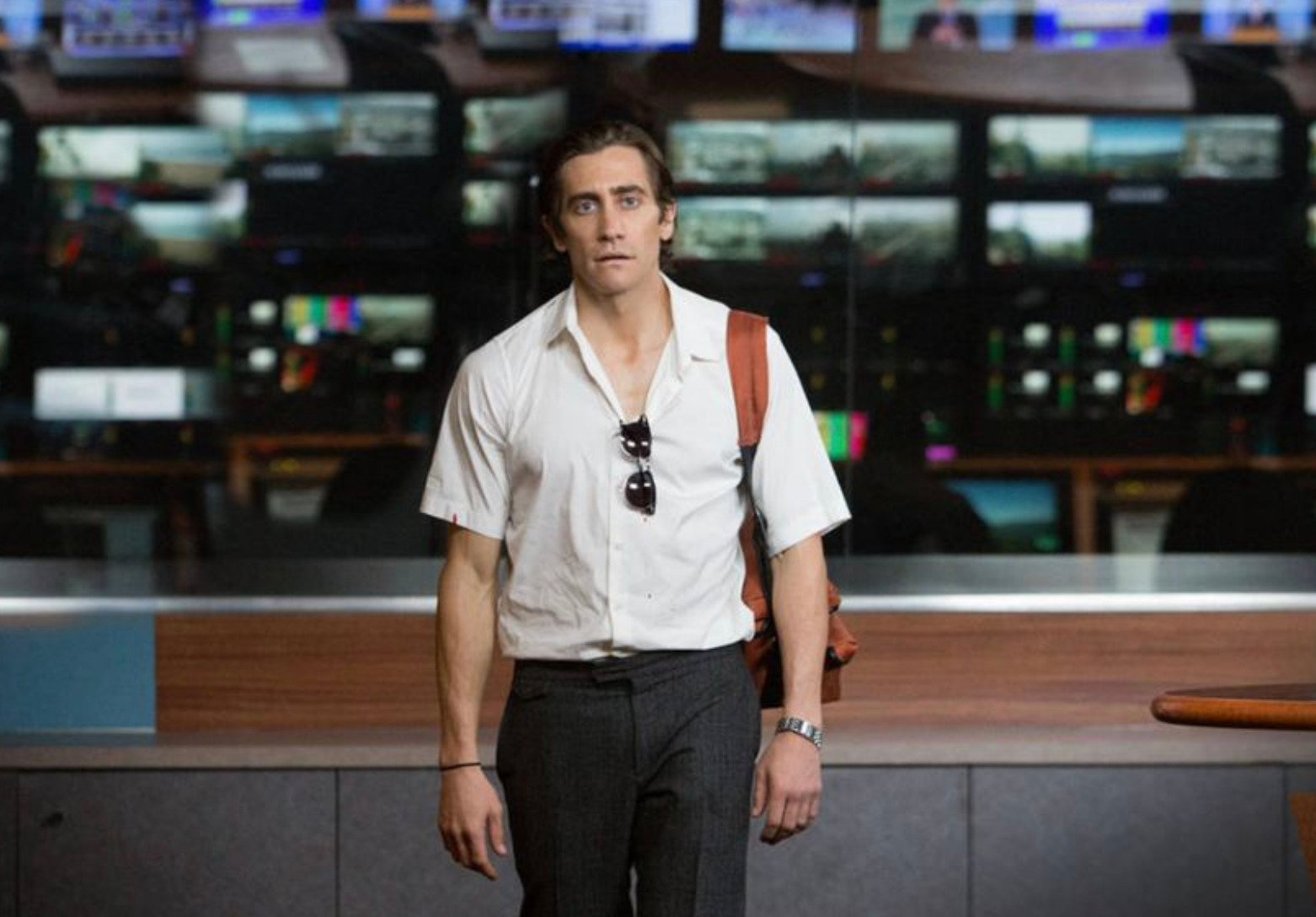 A man with tousled hair, wearing a white short-sleeve shirt, dark pants, sunglasses hanging from his shirt, and a brown bag over his shoulder, stands indoors in front of a wall of TV monitors.