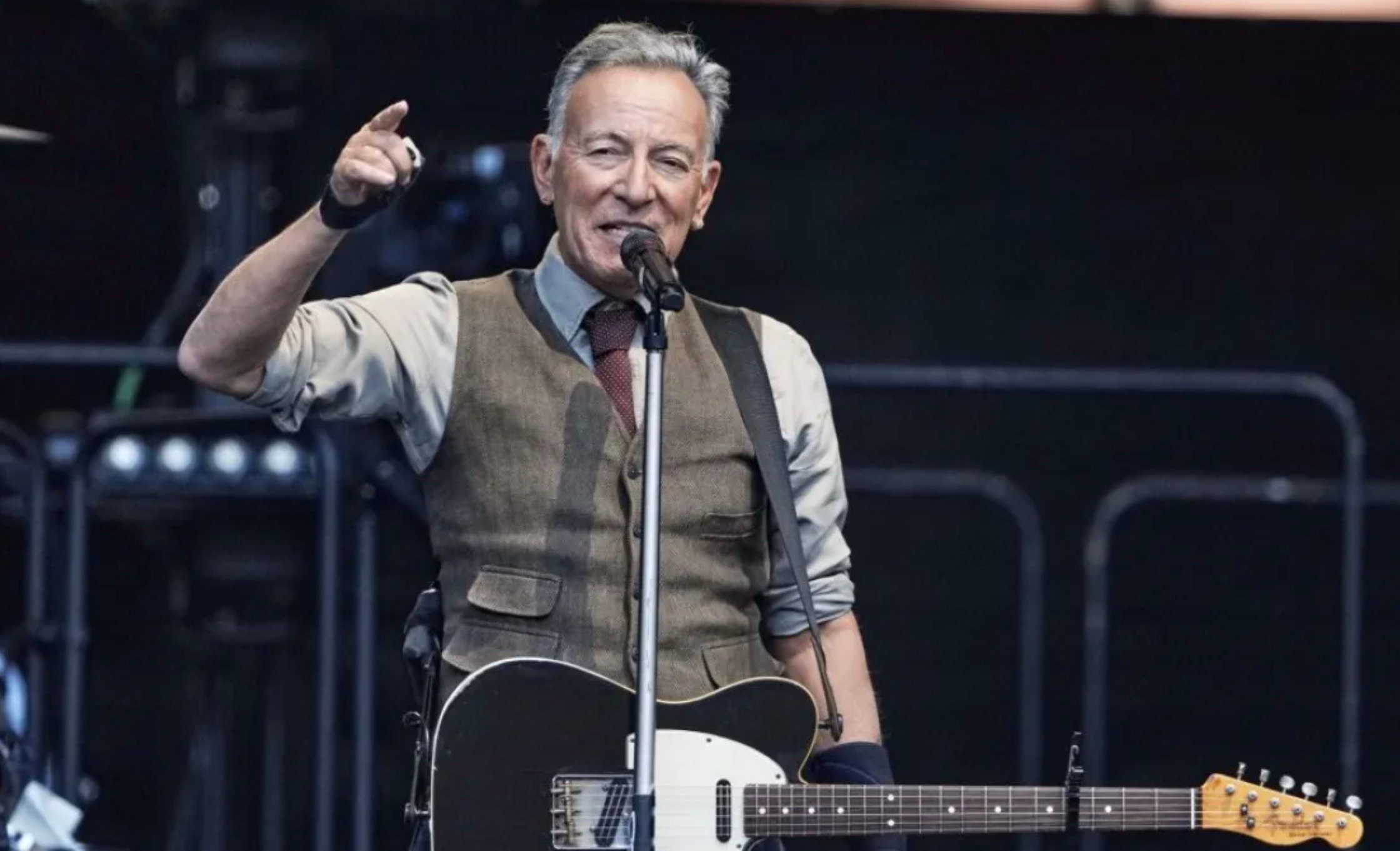 A man wearing a vest and tie stands on stage, holding an electric guitar and singing into a microphone while pointing towards the audience.