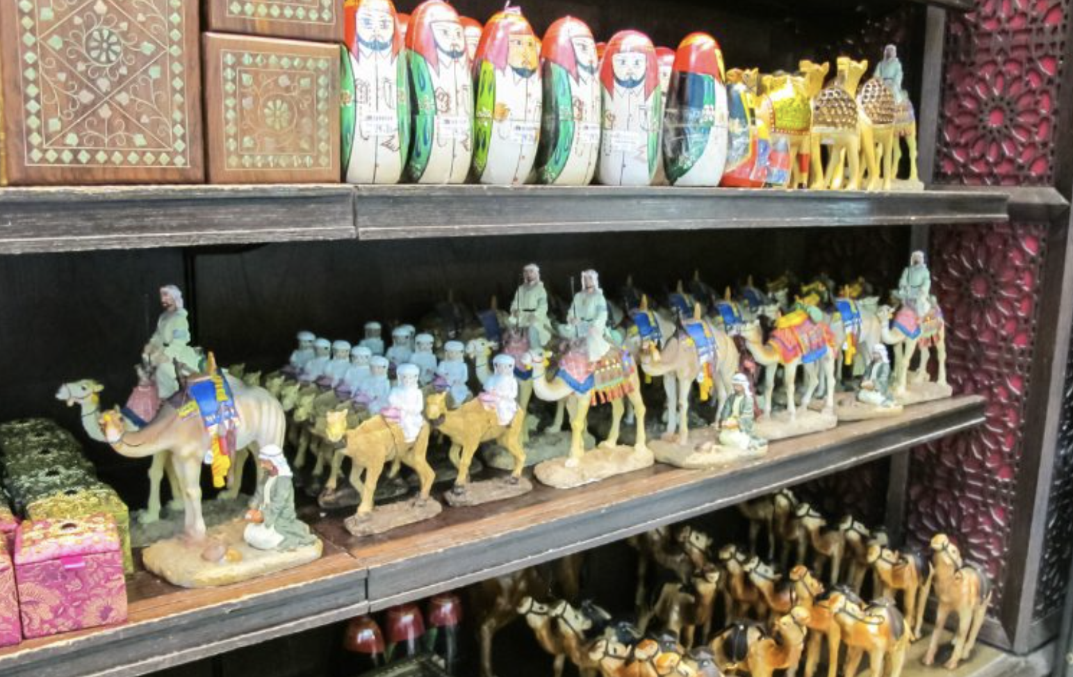 Shelves display colorful nesting dolls, figurines of people riding camels, miniature camels, and decorated boxes with intricate patterns. The items are arranged in neat rows, creating a vibrant and ornate scene.