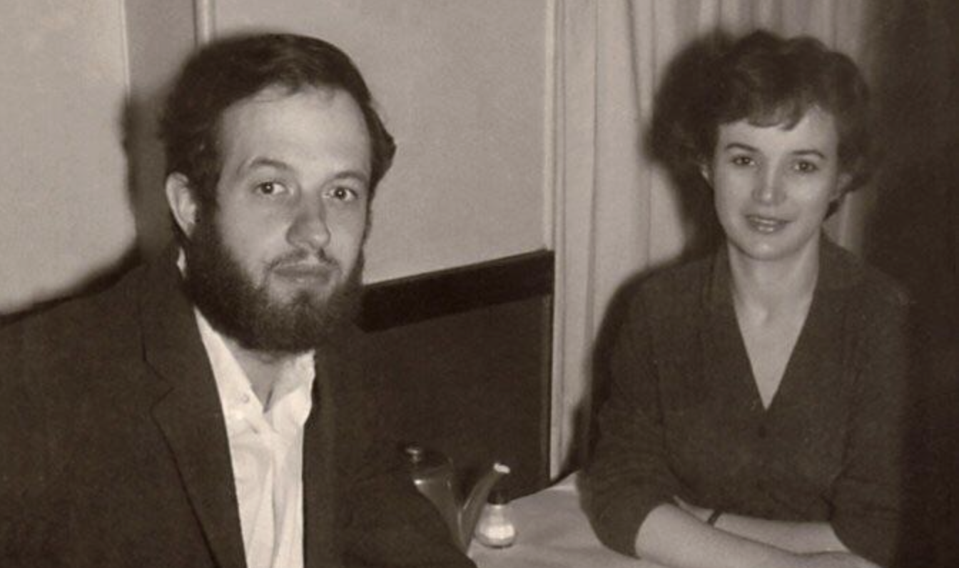 A black-and-white photo of a man with a beard and a woman with short hair sitting at a table together, both looking toward the camera and smiling slightly. A teapot and salt shaker are on the table.