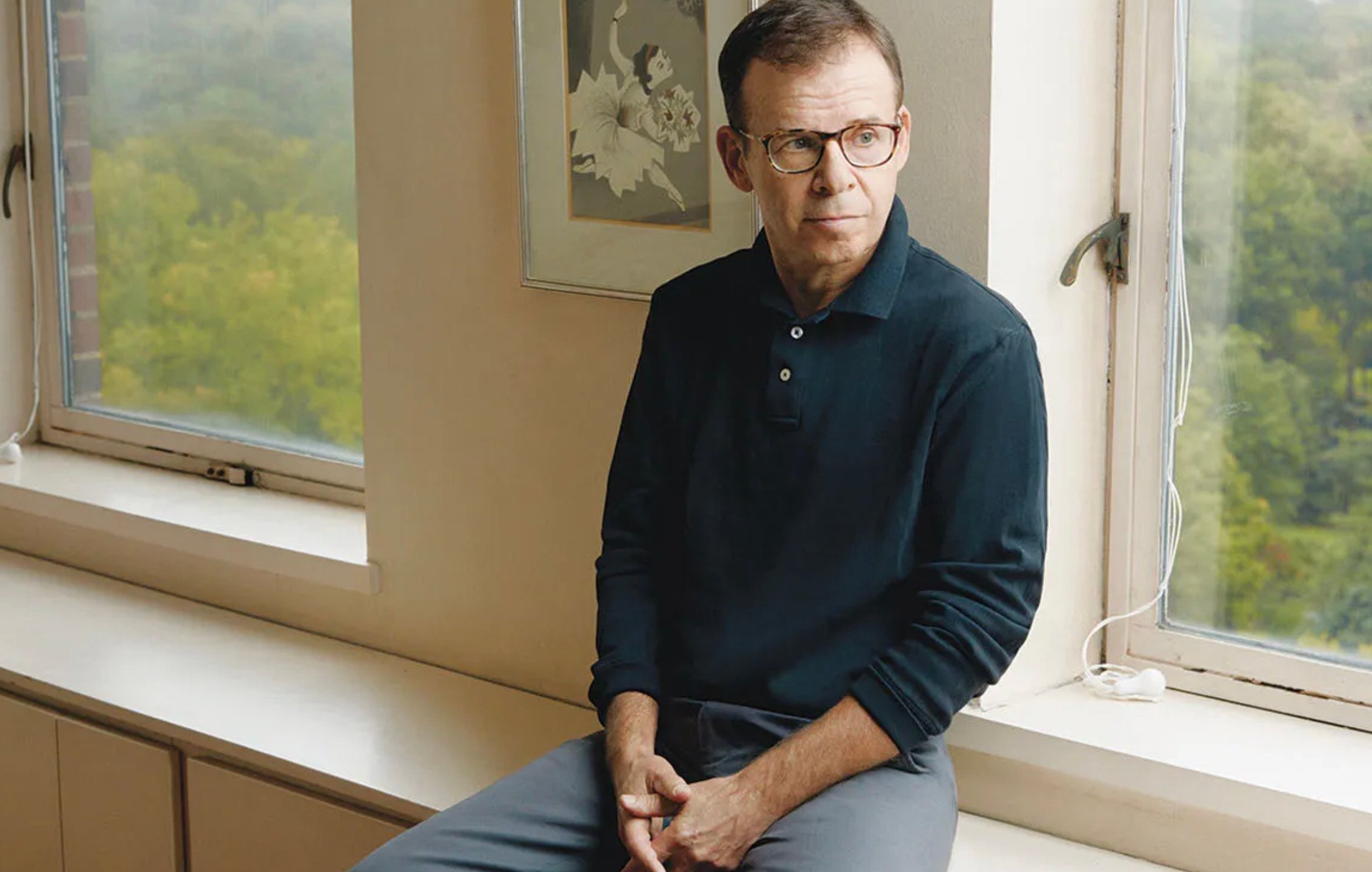 A man wearing glasses and a dark long-sleeve shirt sits on a windowsill, looking thoughtfully to the side. Large windows behind him reveal a view of trees outside. A framed artwork hangs on the wall.