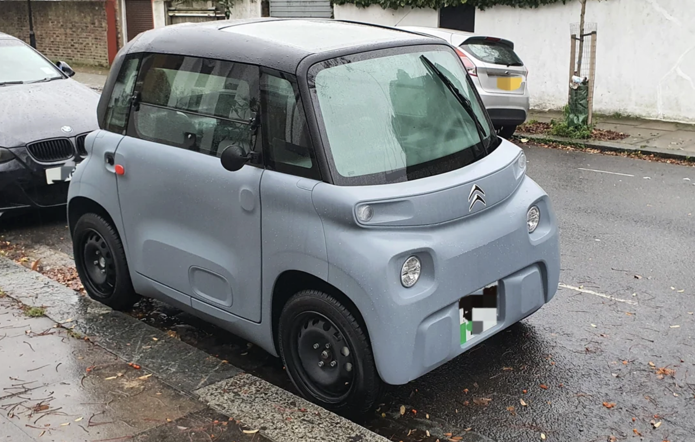 A small, light blue Citroën Ami electric car is parked on a wet street beside a curb, with other cars nearby and fallen leaves scattered on the ground.