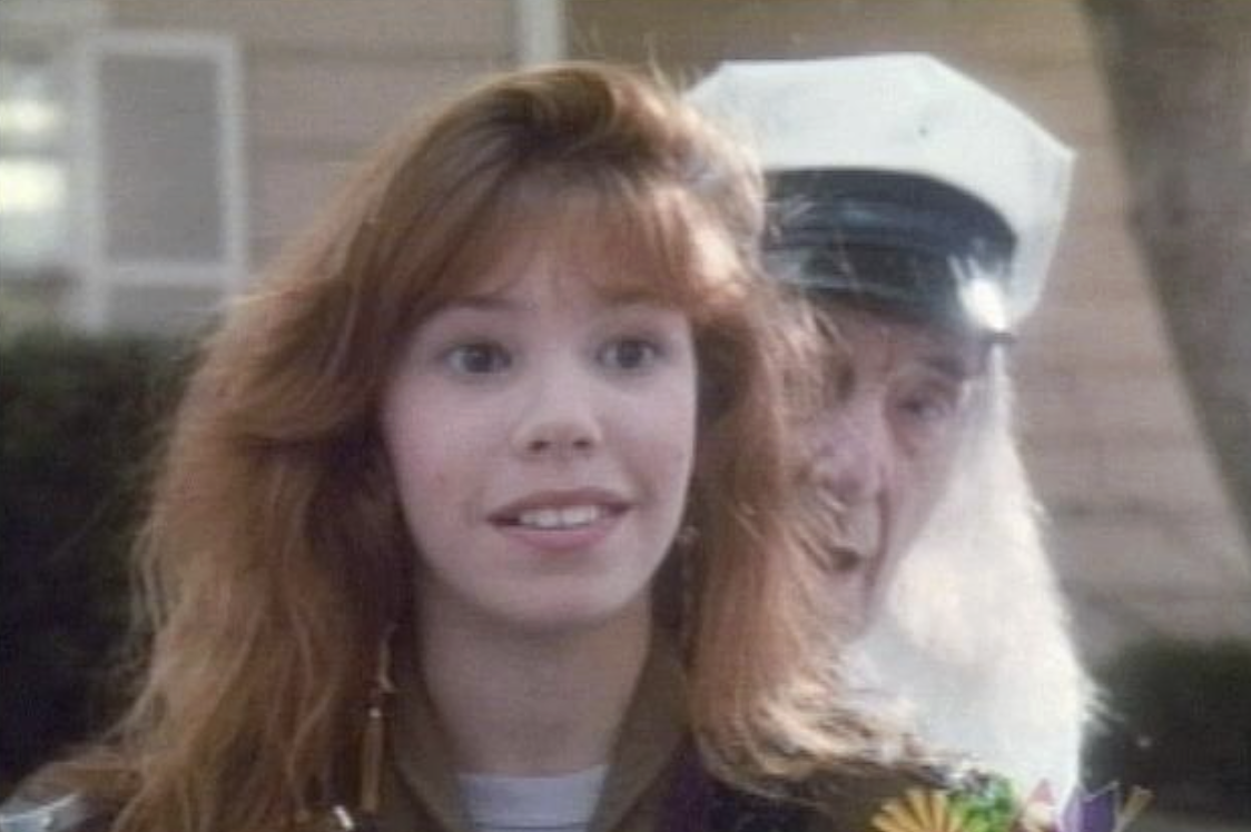 A young woman with long reddish hair stands outside, smiling slightly. An older person in a white hat and uniform appears behind her, partially obscured and looking in her direction.