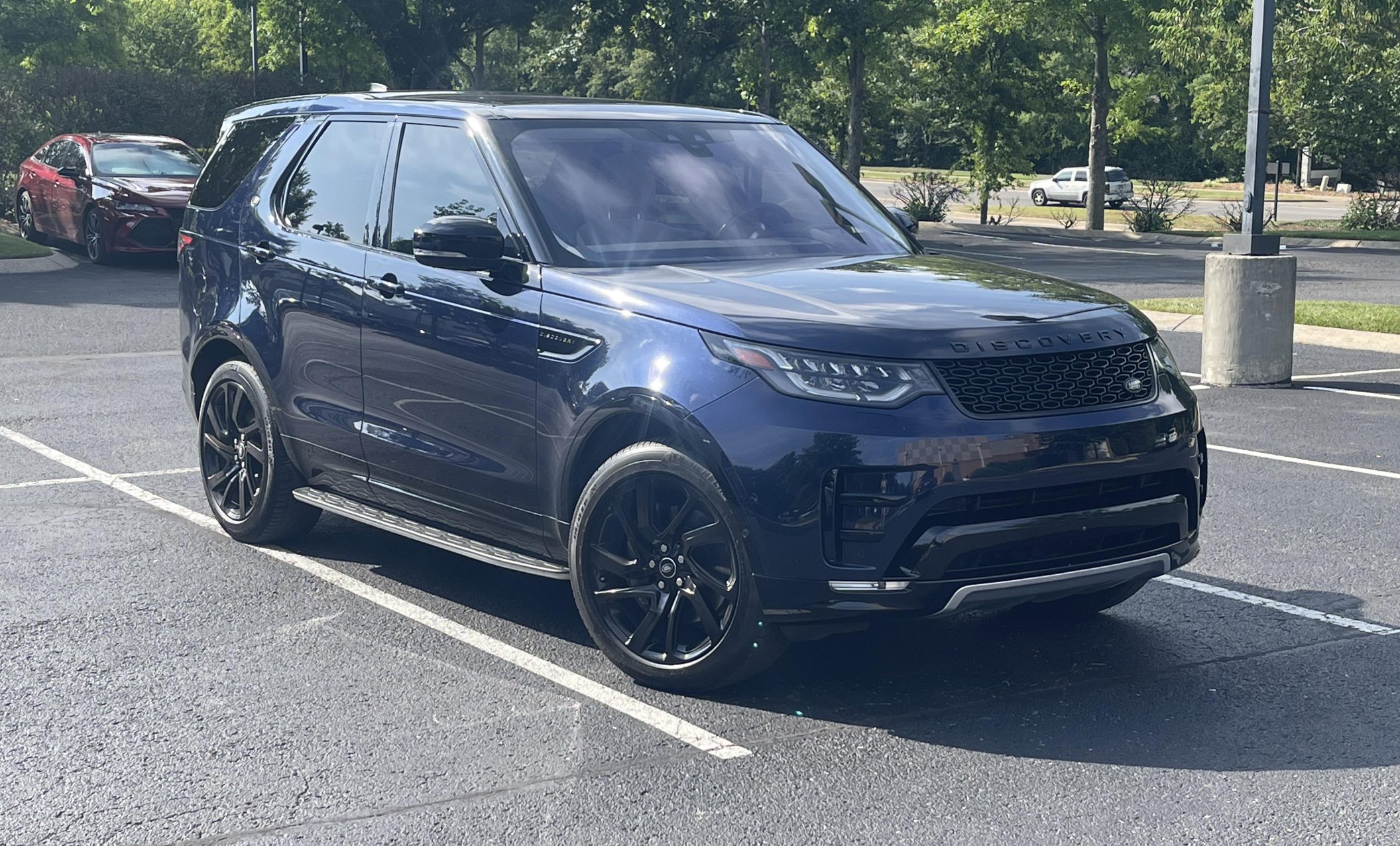 A dark blue Land Rover Discovery SUV with tinted windows is parked in a sunny parking lot, surrounded by trees and a few other cars in the background.