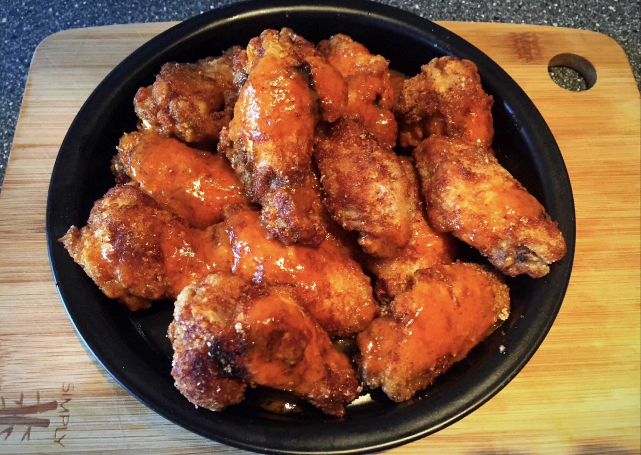 A black plate piled high with crispy chicken wings coated in a shiny orange sauce, placed on a wooden cutting board with a tree logo. The background surface is a speckled countertop.