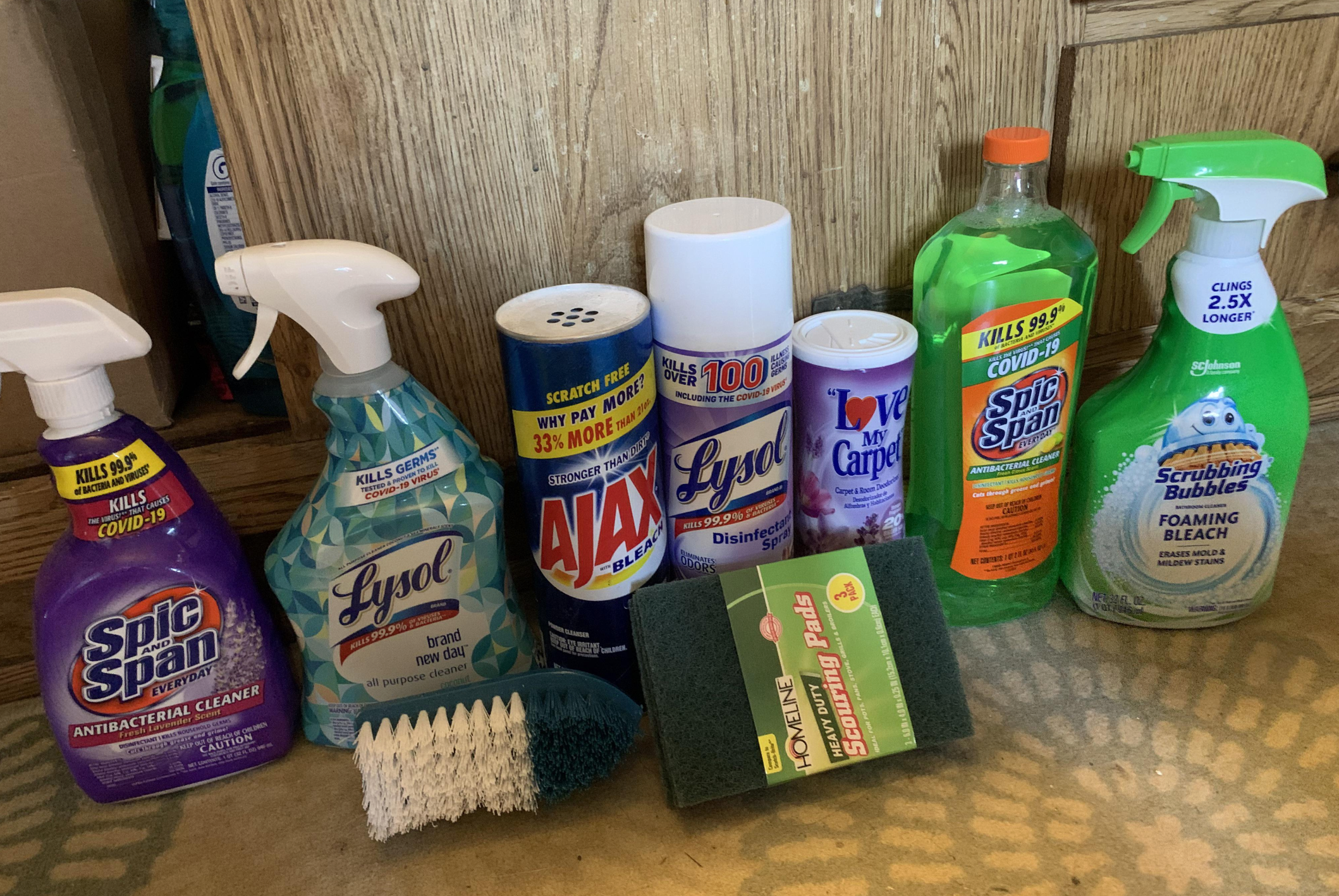 A variety of cleaning products, including Spic and Span, Lysol, Ajax powder, a scrub brush, Love My Carpet spray, Scrubbing Bubbles, a green sponge, and a bottle of dish soap, are arranged on a carpeted floor.