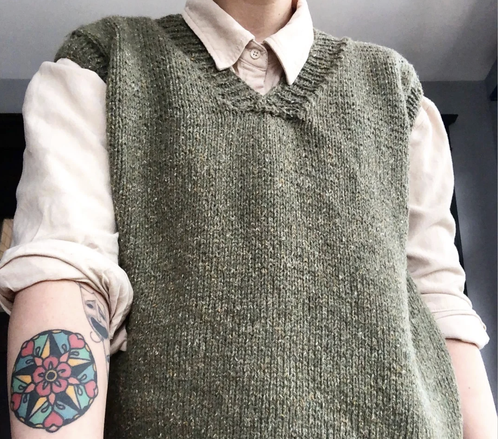 Person wearing a light beige collared shirt with rolled-up sleeves and an olive green knitted vest; a colorful mandala tattoo is visible on their left forearm.