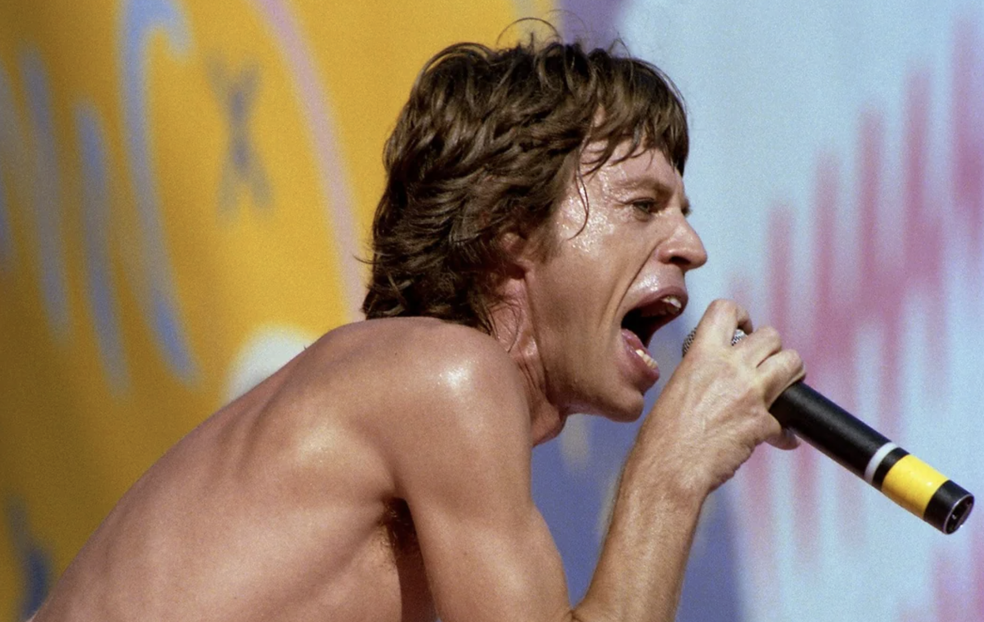 A shirtless man with sweaty skin and messy brown hair passionately sings into a microphone against a colorful, blurred background.