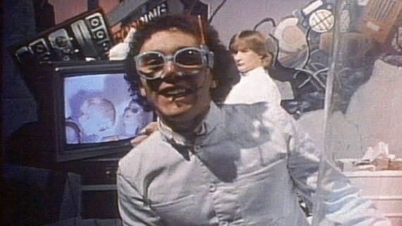 A person wearing large, quirky goggles and a white outfit smiles at the camera. Another person stands behind them. The background features a TV with a face on the screen and colorful, chaotic props.