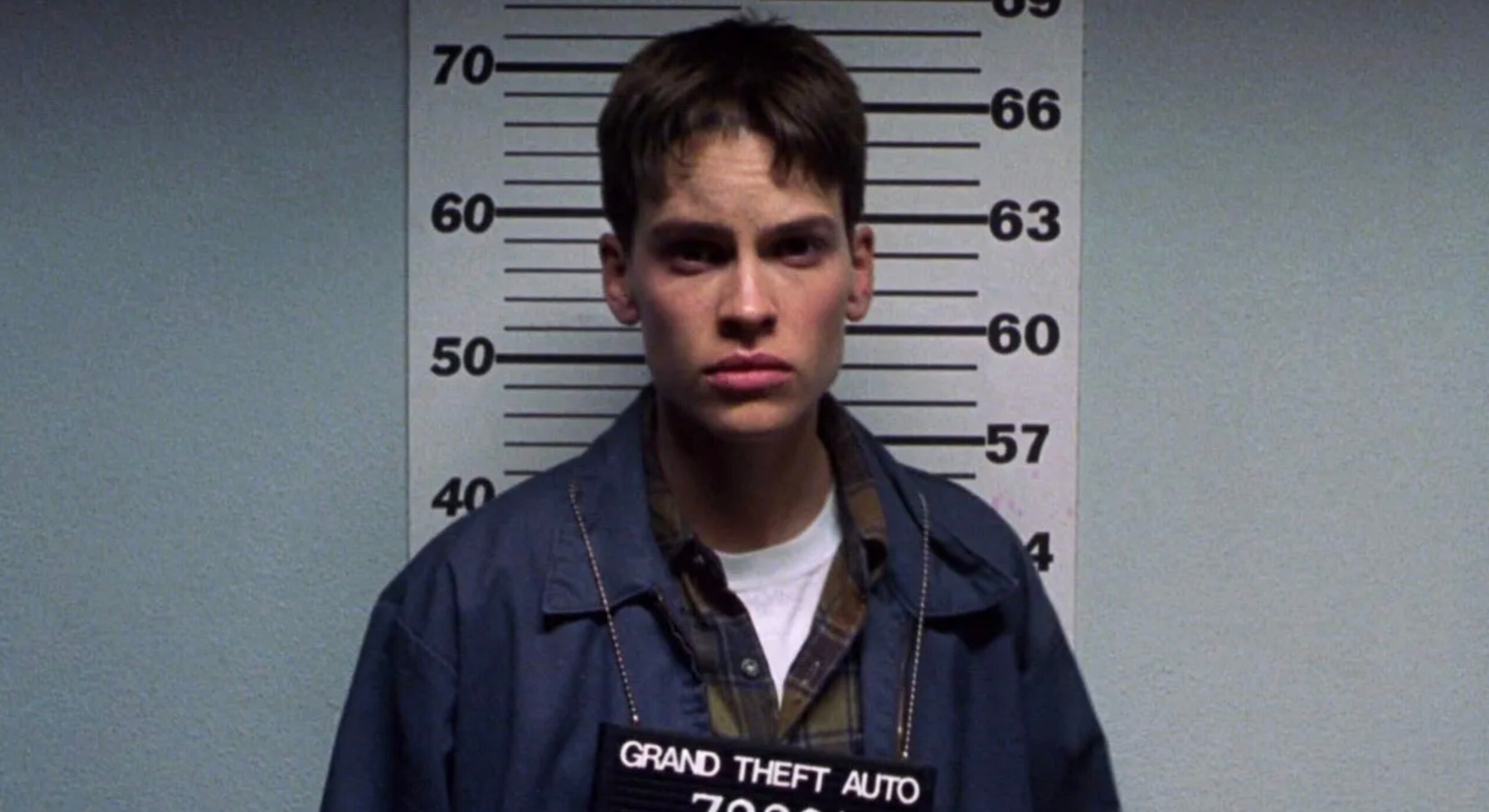 A person with short hair stands in front of a height chart holding a sign that reads "Grand Theft Auto 72031," wearing a jacket over a plaid shirt, with a serious expression.
