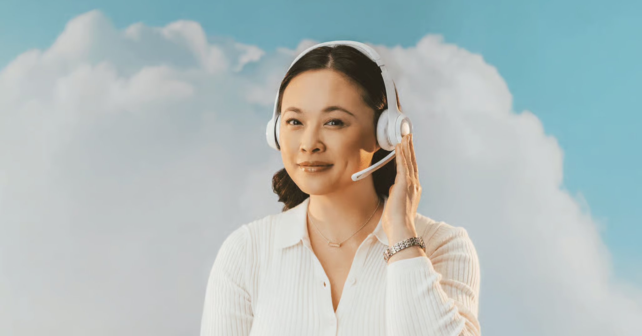 A woman wearing a white headset and white top smiles while touching her headset, standing in front of a backdrop featuring fluffy white clouds and a blue sky.
