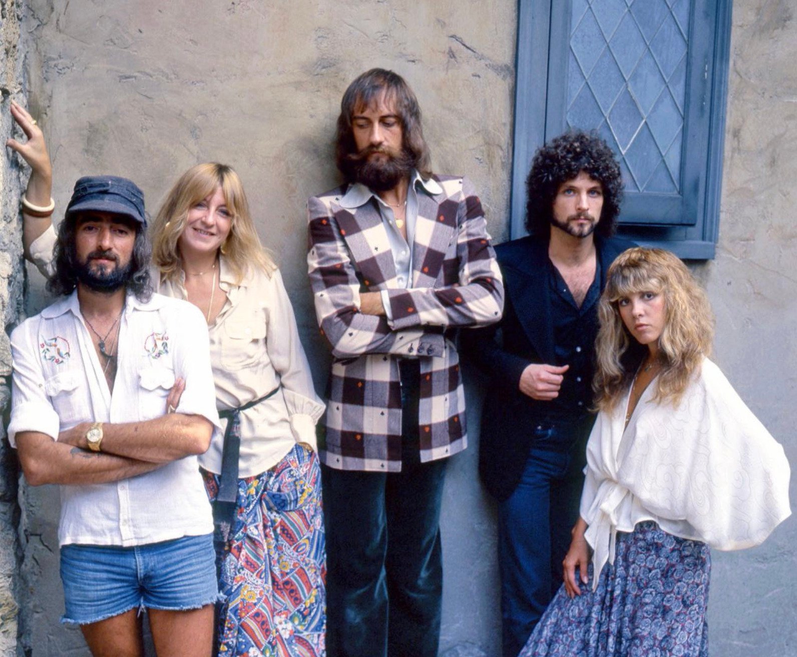 Five people stand against a stone wall, posing casually. Three men and two women wear 1970s-style clothing, including a plaid jacket, skirt, shorts, and flowing garments. One window is visible behind them.
