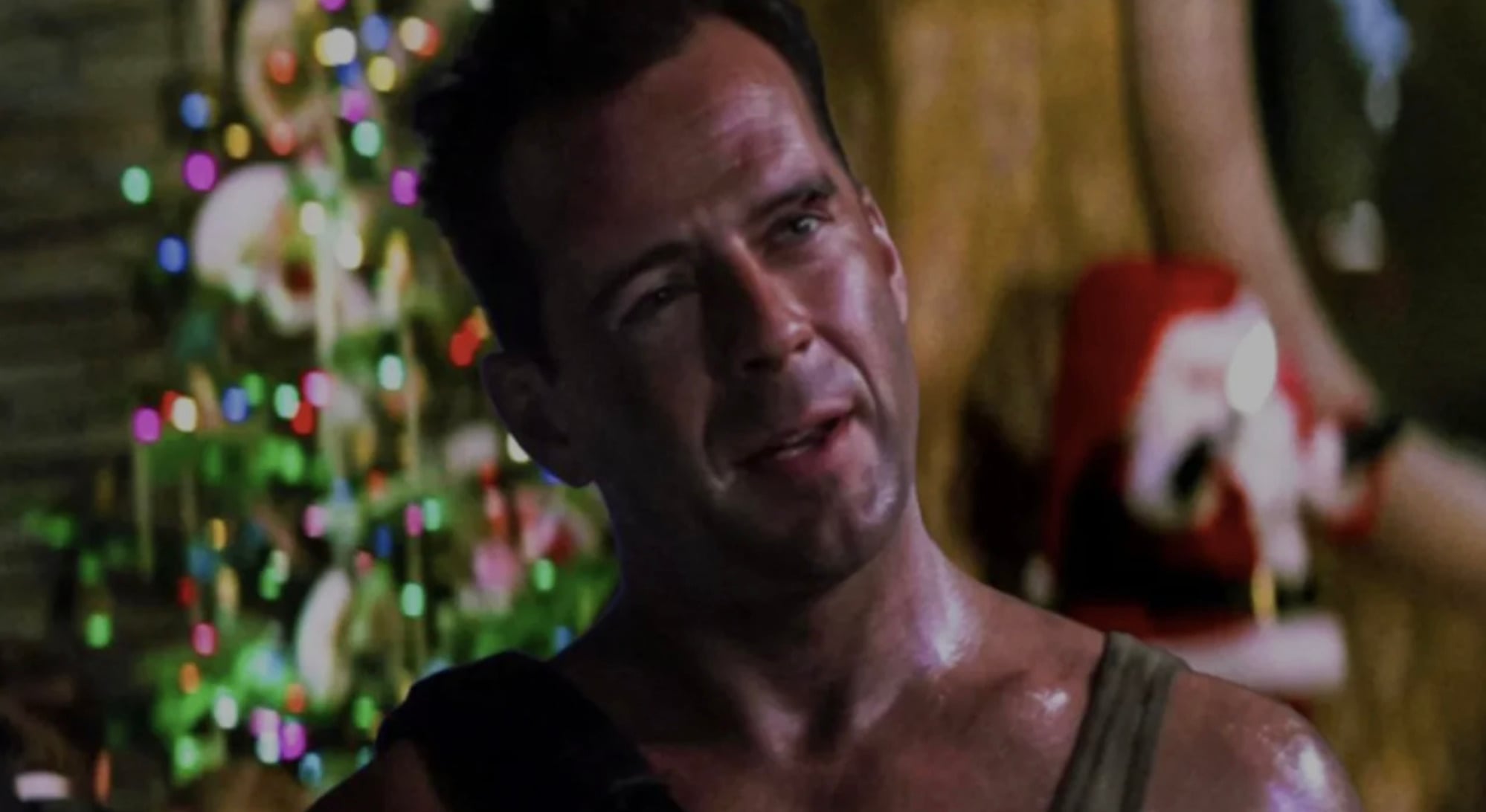 A man with short hair, wearing a tank top and looking slightly to the side, stands in front of a decorated Christmas tree with colorful lights in a dimly lit room.