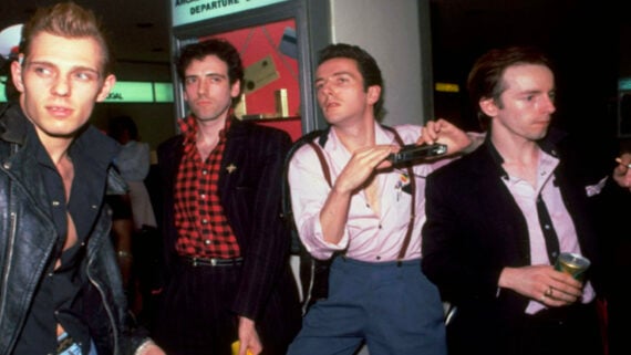 Four men in stylish 1980s attire stand together indoors; one holds a camera, another a drink. A departure board is visible behind them, suggesting they are at an airport. The mood appears candid and energetic.