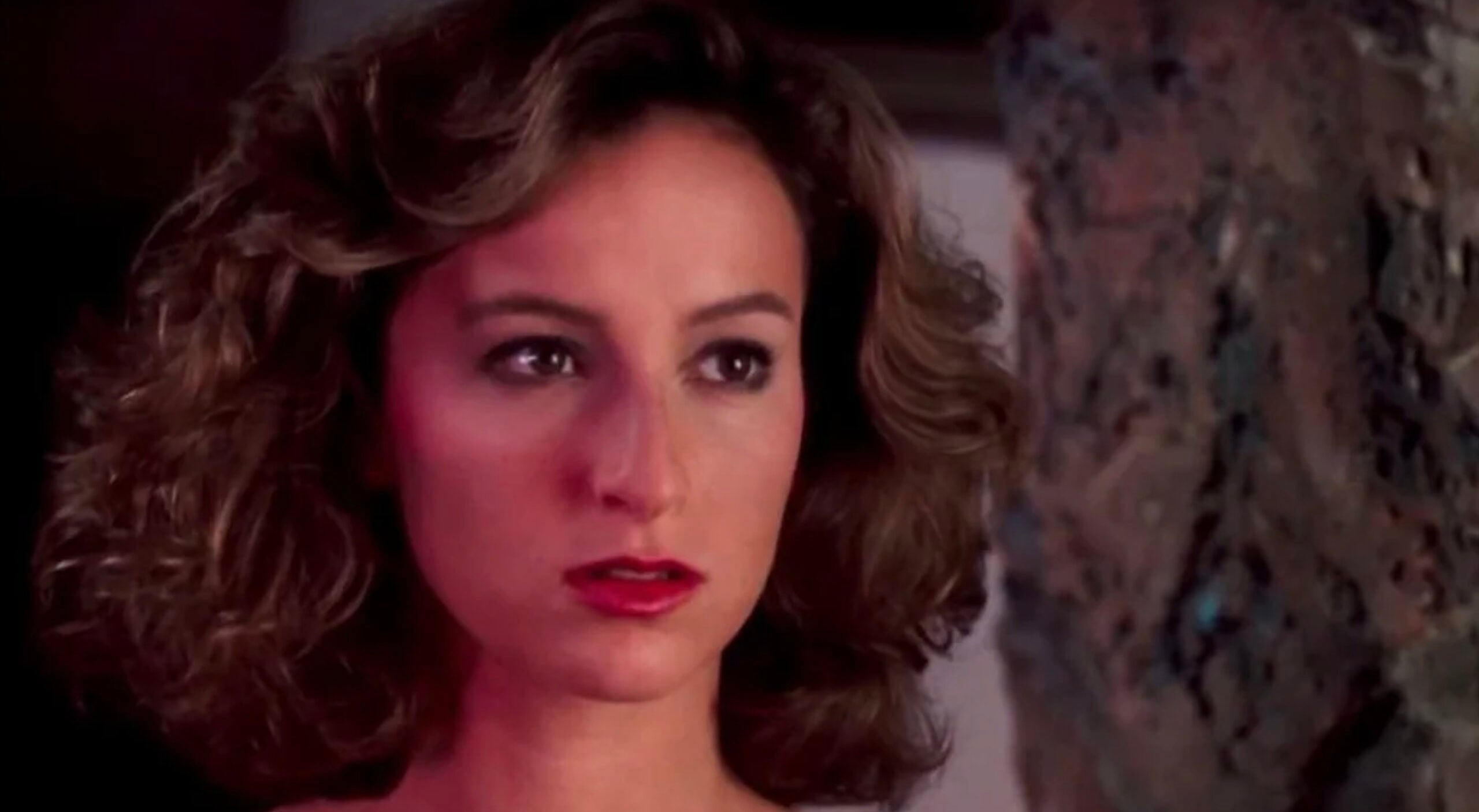 A woman with curly brown hair and red lipstick looks intently to the side, her expression serious, with a blurred, textured object in the foreground on the right.