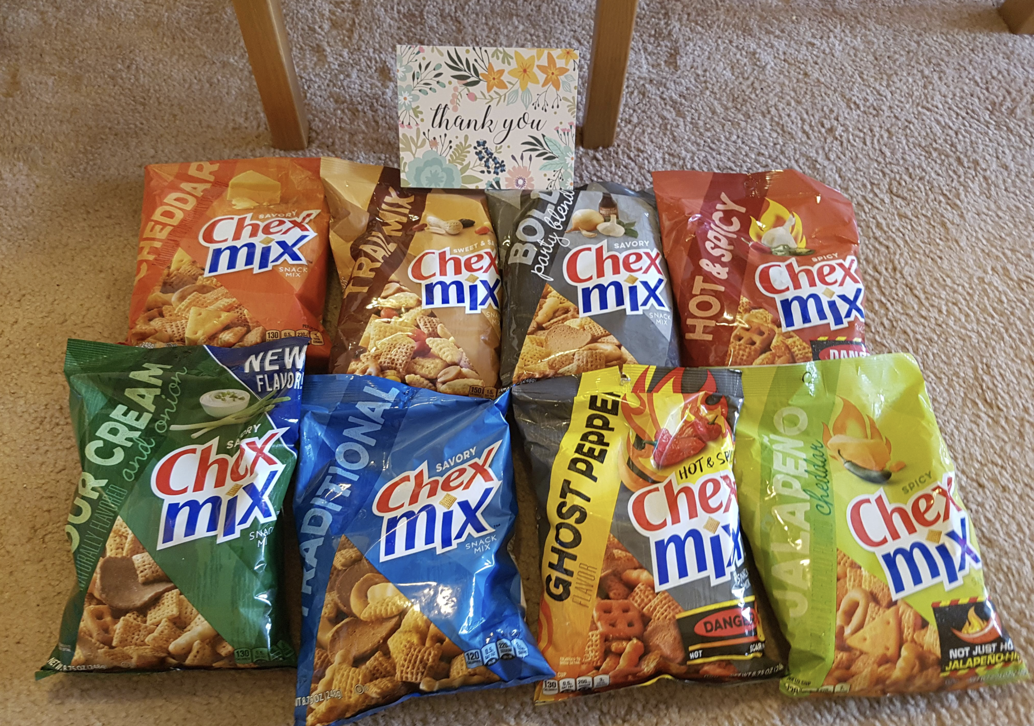Nine assorted bags of Chex Mix snacks are arranged on a beige carpet with a “thank you” card propped up behind them on a wooden chair leg. The flavors include Cheddar, Traditional, Honey BBQ, Hot & Spicy, Sour Cream & Onion, Ghost Pepper, and Jalapeno.