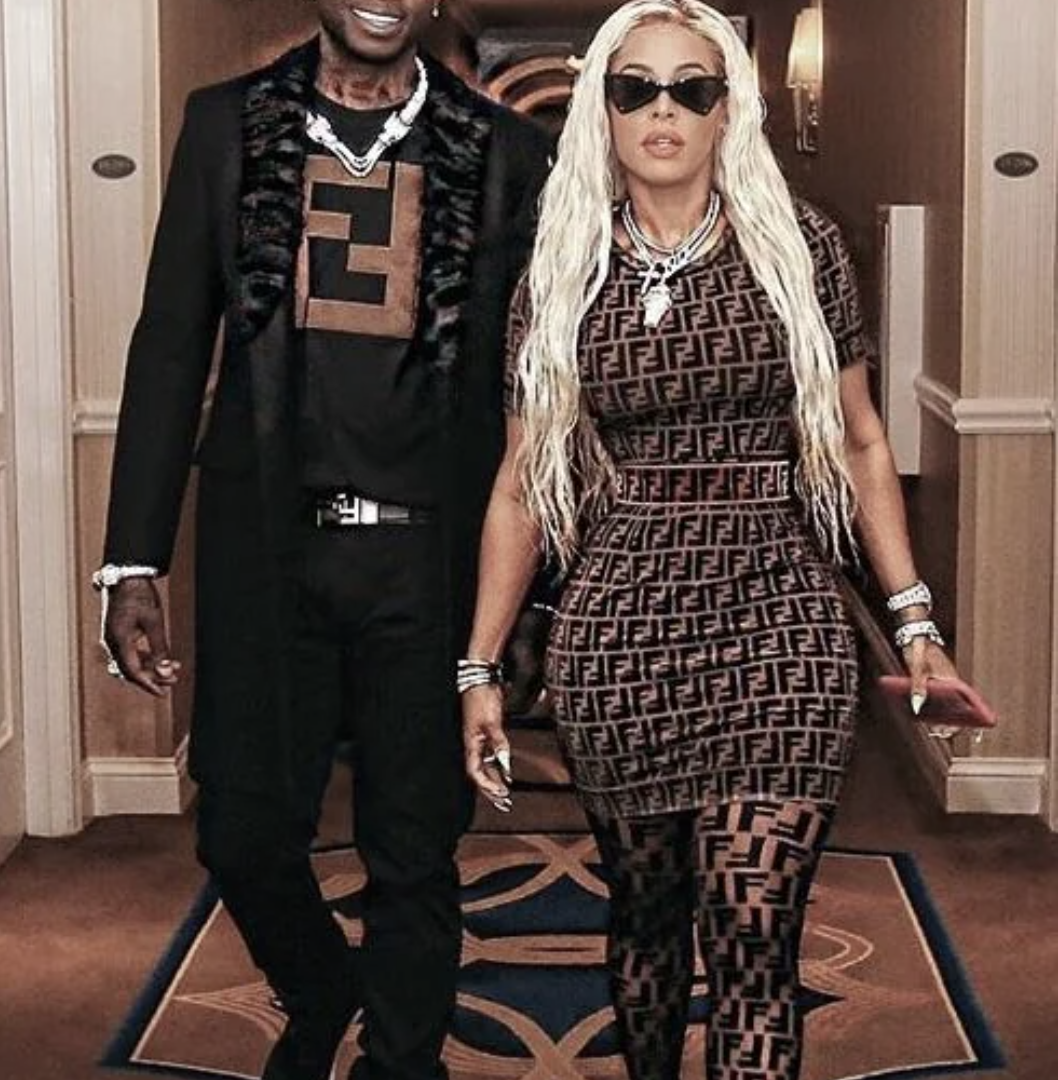 A man and a woman walk down a hallway wearing matching black and brown Fendi outfits and jewelry. The woman has long blonde hair, sunglasses, and a fitted dress, while the man wears a coat and a T-shirt with the Fendi logo.