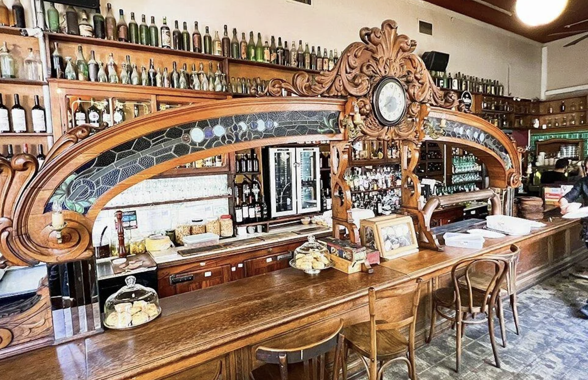 A vintage wooden bar with ornate carvings, glass shelves filled with bottles, pastries on display, framed photos, and wooden chairs, creating a classic and cozy atmosphere.