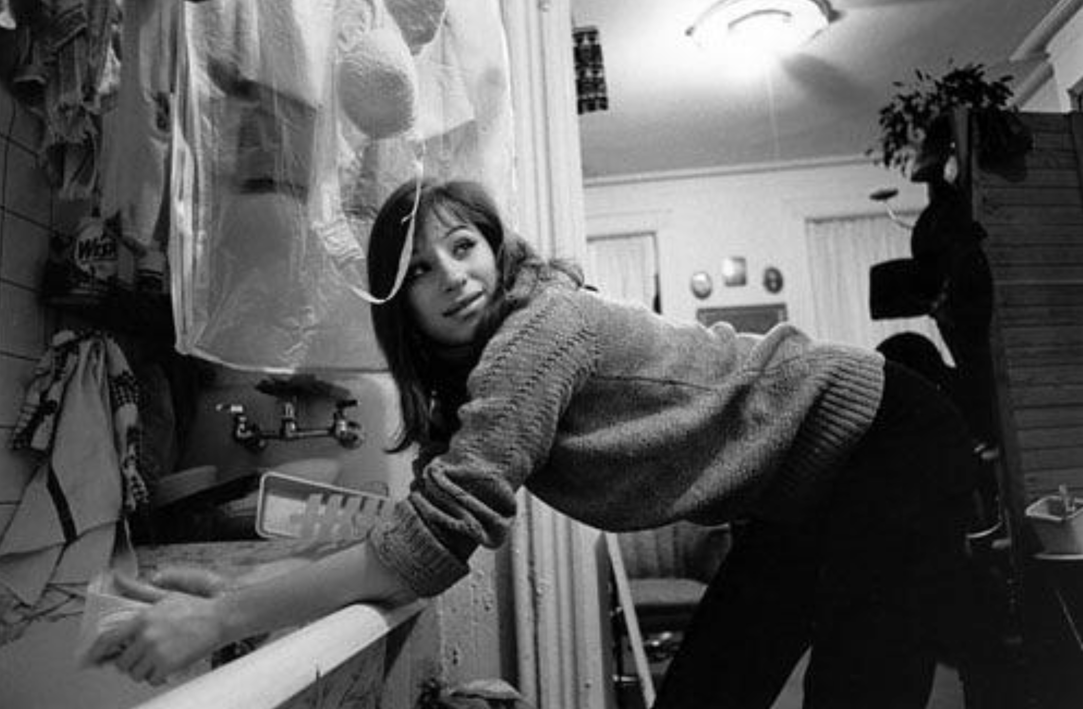 A woman in a sweater washes dishes at a kitchen sink, glancing over her shoulder and smiling. The kitchen is cluttered, and a bright ceiling light illuminates the scene.