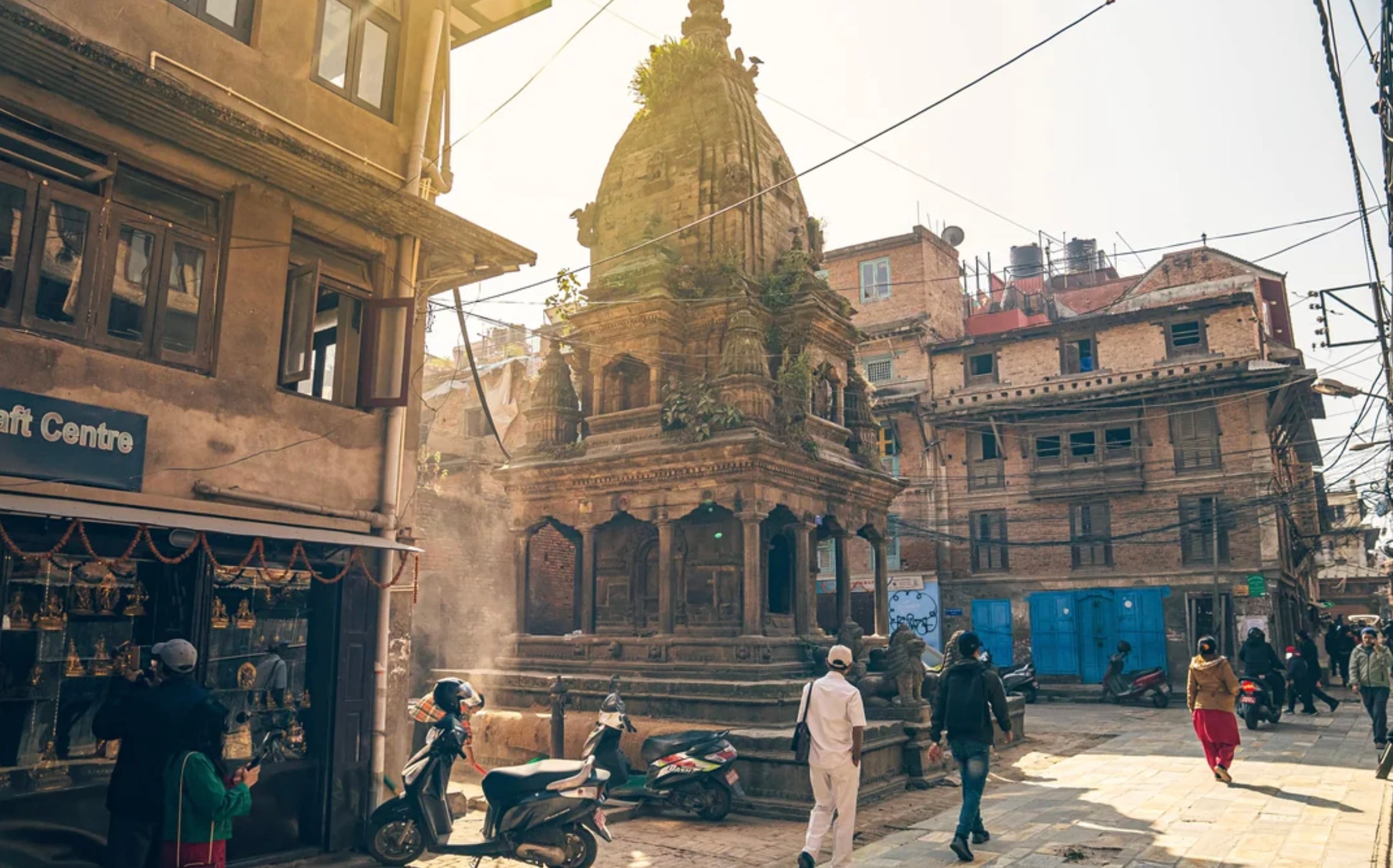 People walk and ride scooters near an old stone temple surrounded by traditional buildings in a sunlit street, with prayer wheels and wires visible in the scene.