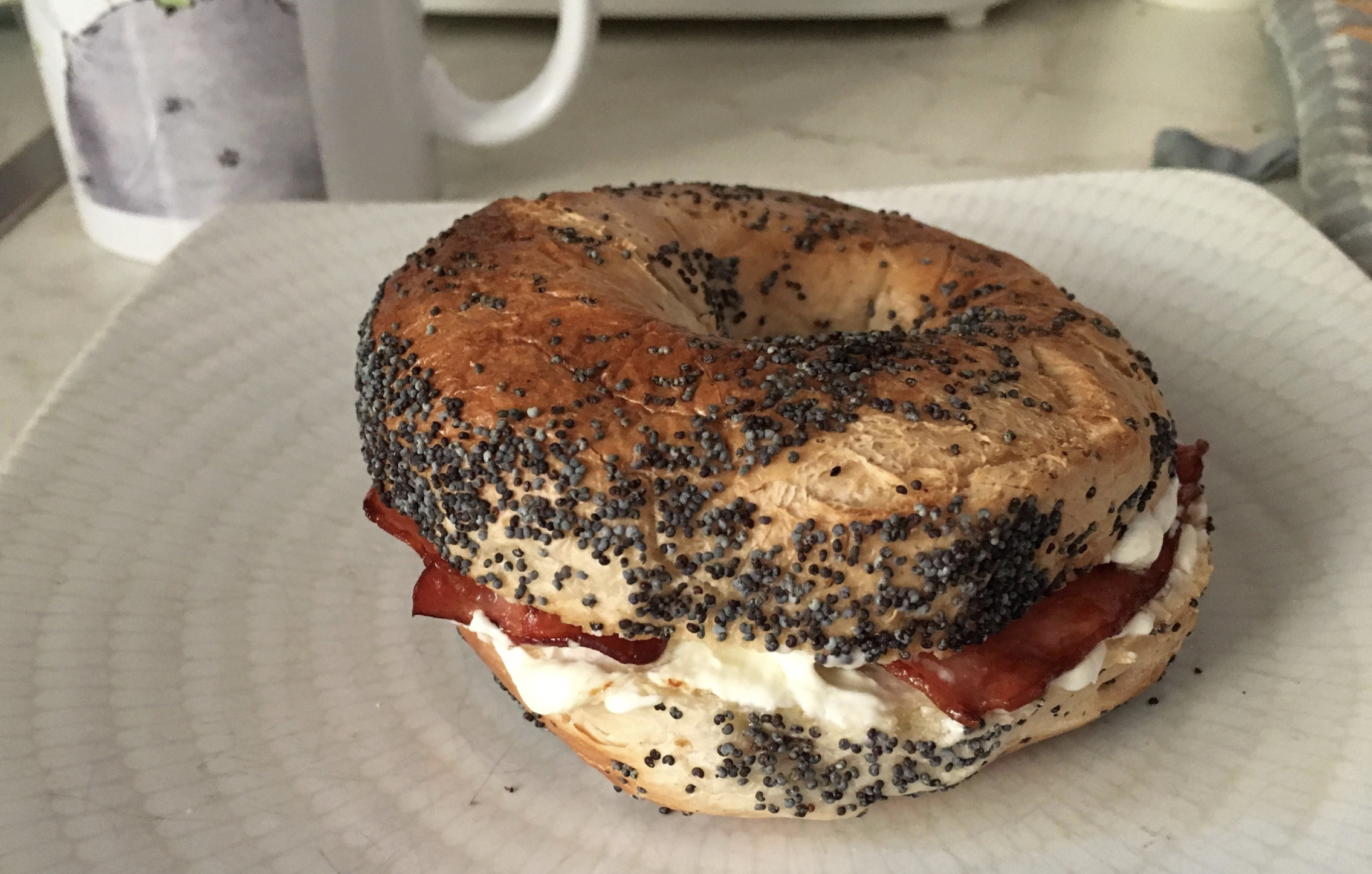 A poppy seed bagel sandwich with cream cheese and bacon sits on a white plate, with a mug partially visible in the background.