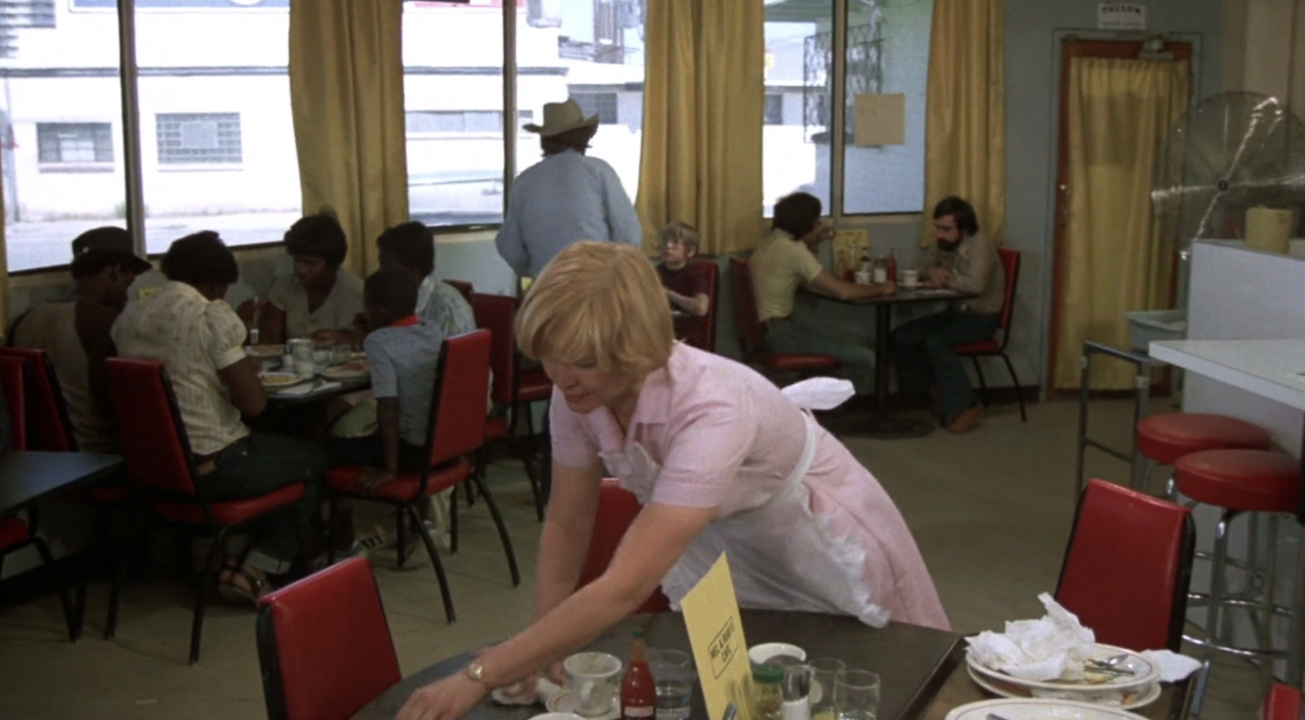 A waitress in a pink uniform clears a table in a casual diner with red chairs and yellow curtains. Several groups of people are seated and eating at tables near the windows.