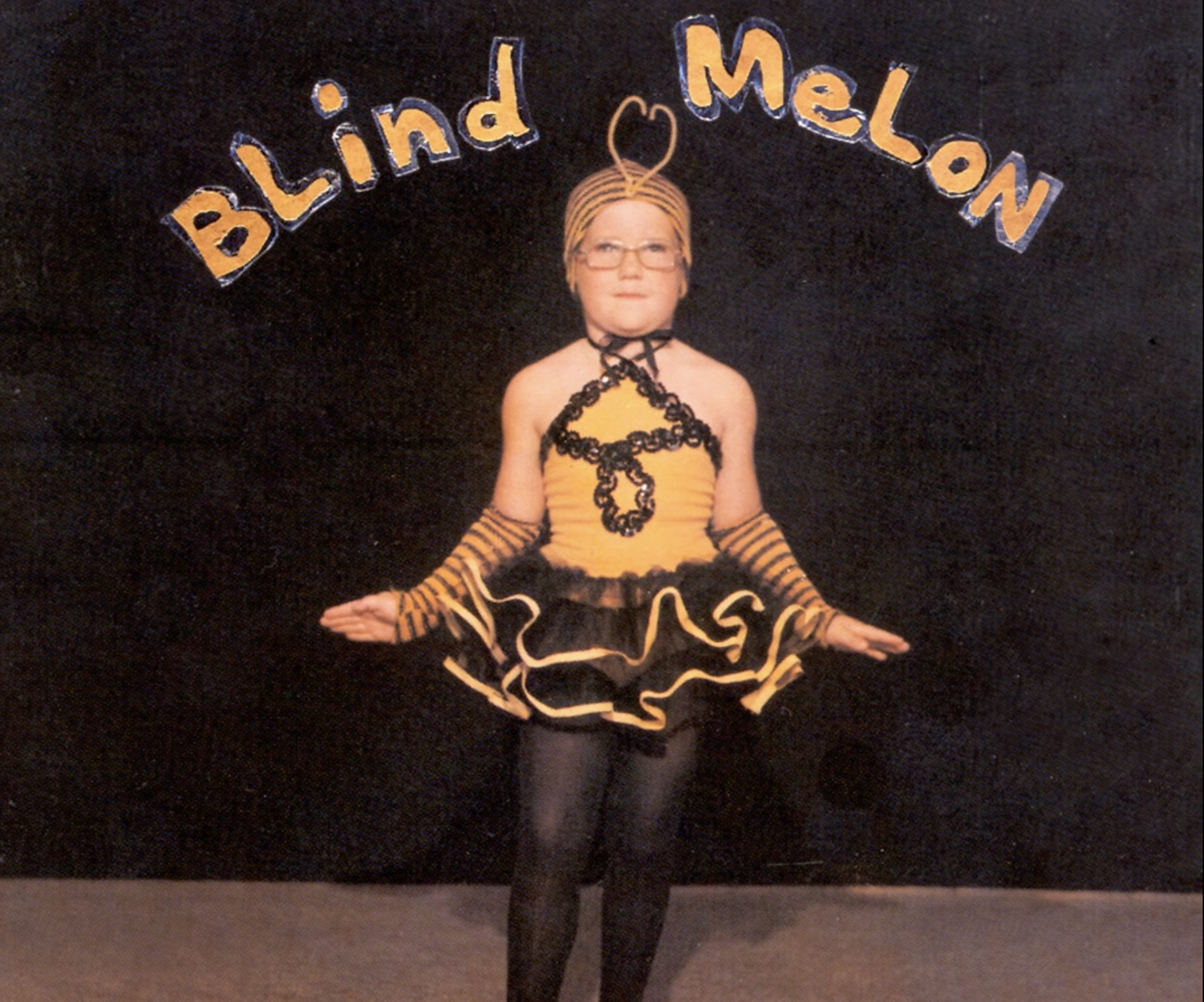 A child in a bee costume stands on stage against a dark background, with "Blind Melon" written in yellow, stylized text above them.