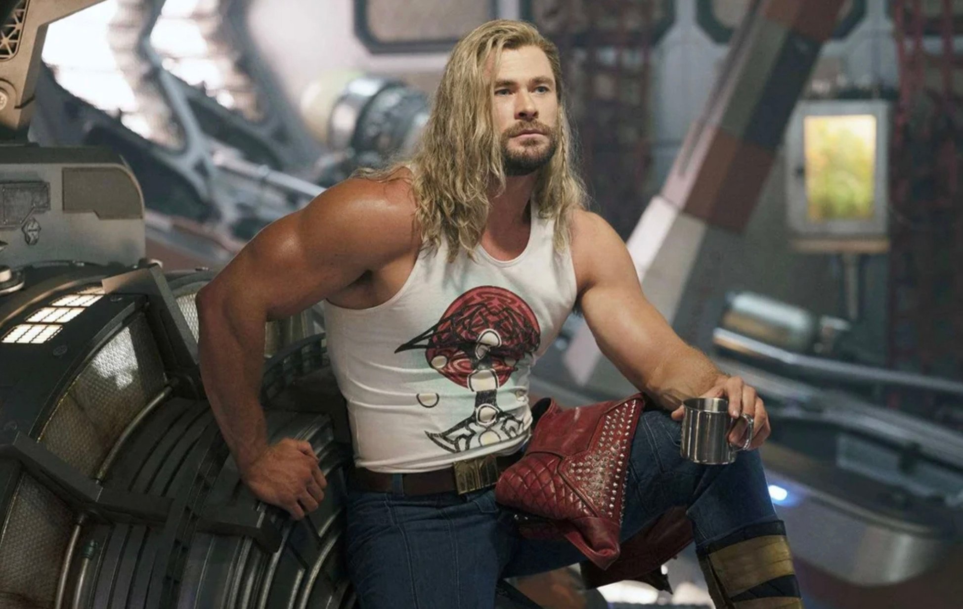 A muscular man with long blond hair sits on a metal structure in a sci-fi setting, wearing a white tank top with a graphic, blue jeans, and holding a silver mug. He appears deep in thought.