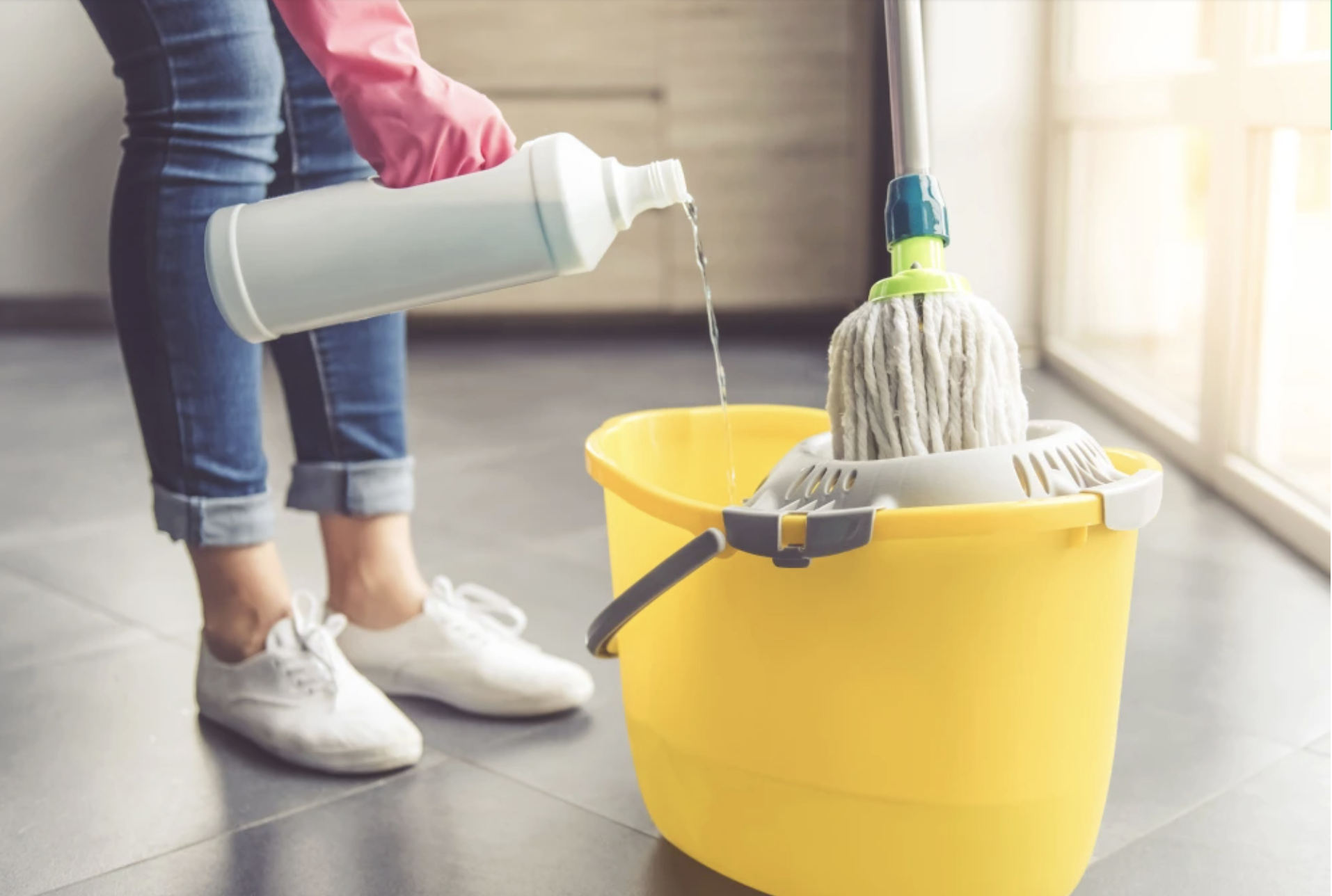 A person wearing jeans, white sneakers, and a pink glove pours cleaning solution from a white bottle into a yellow bucket with a mop, preparing to clean a tiled floor.