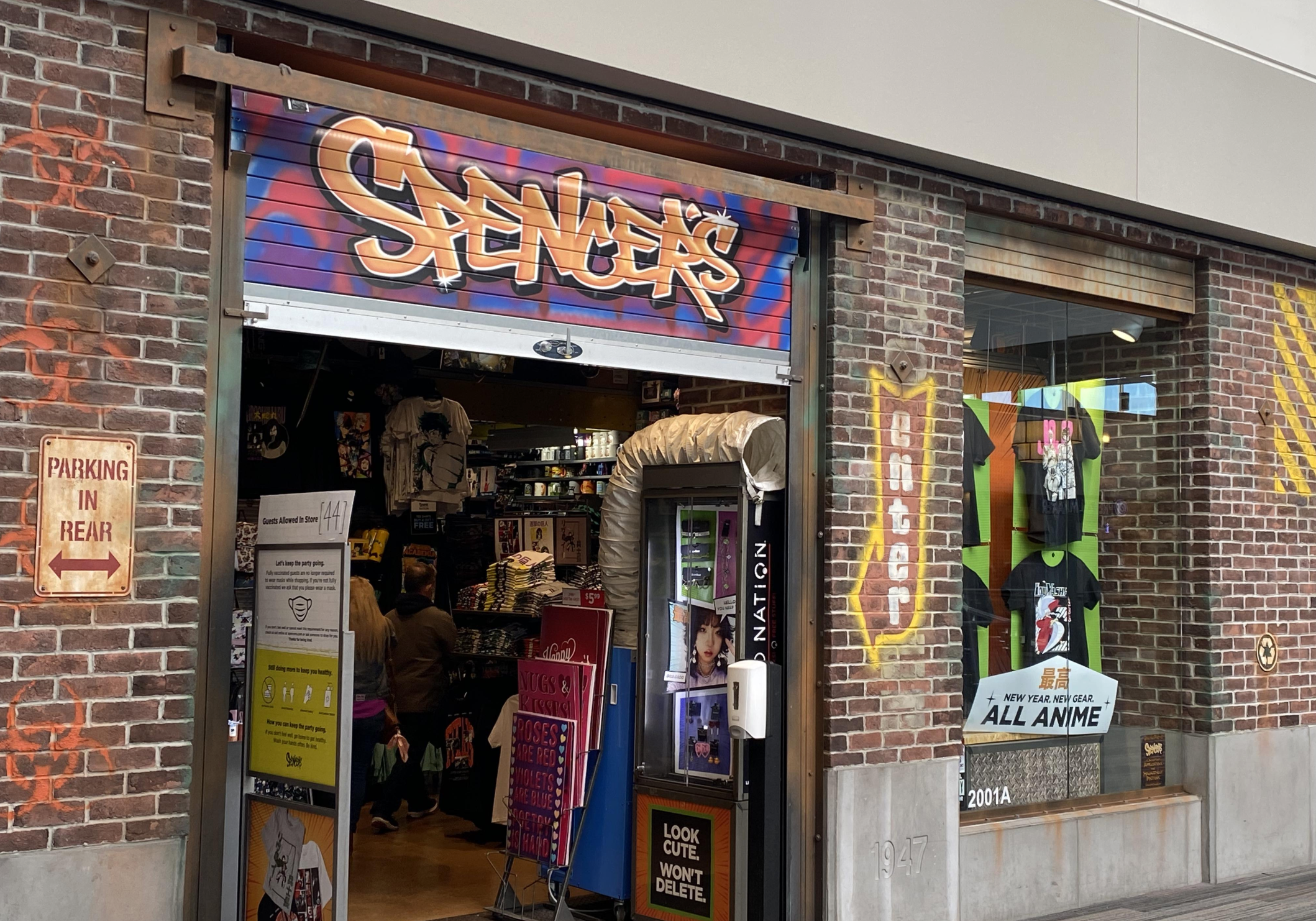 A storefront for Spencer’s in a mall, featuring a colorful sign above the door, T-shirts in the window display, a brick facade, and a "Parking in Rear" sign on the left wall.
