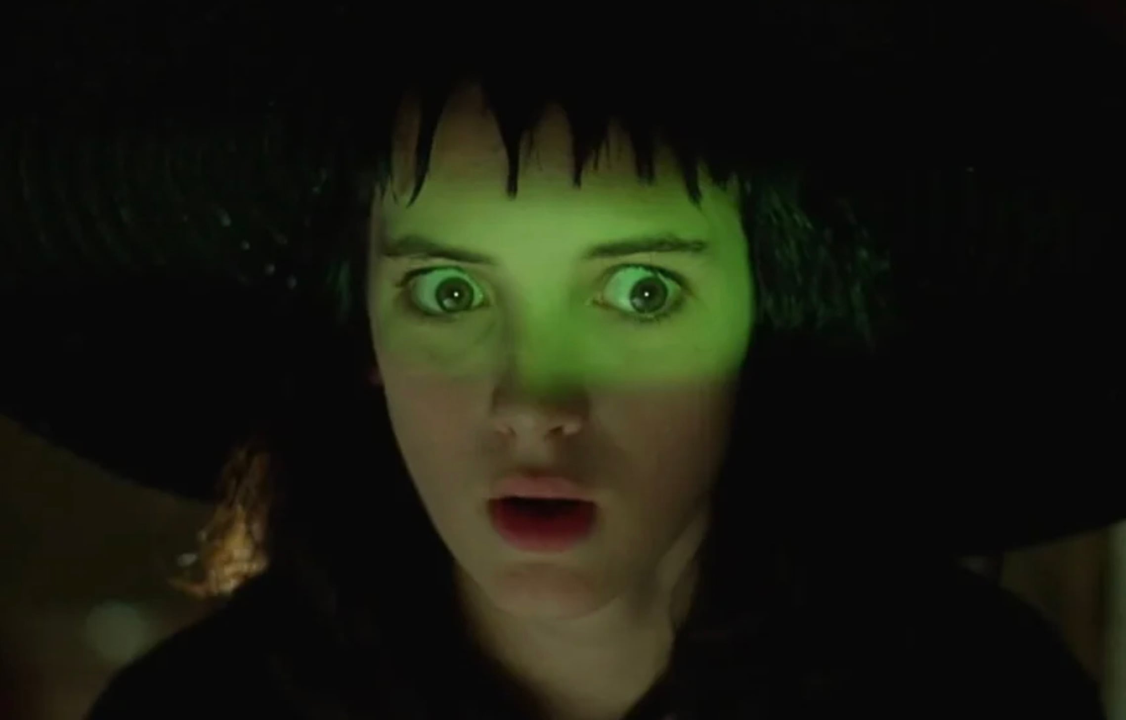 A young woman with wide eyes and parted lips looks surprised or scared. She is lit by green light and wears a black hat with shadows over her face.