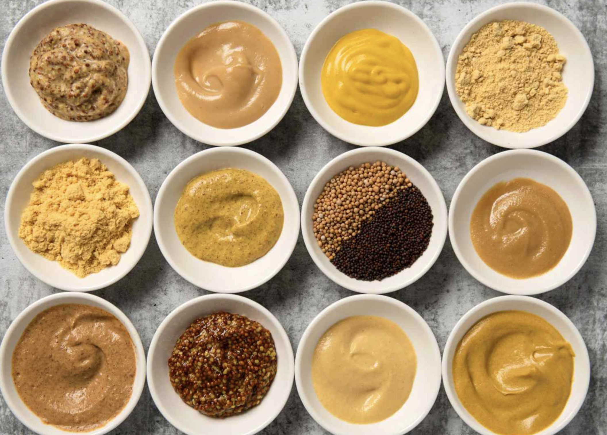 Twelve small white bowls are arranged in a grid, each containing a different type or variety of mustard, including smooth, whole grain, powdered, and coarse textures, on a gray surface.