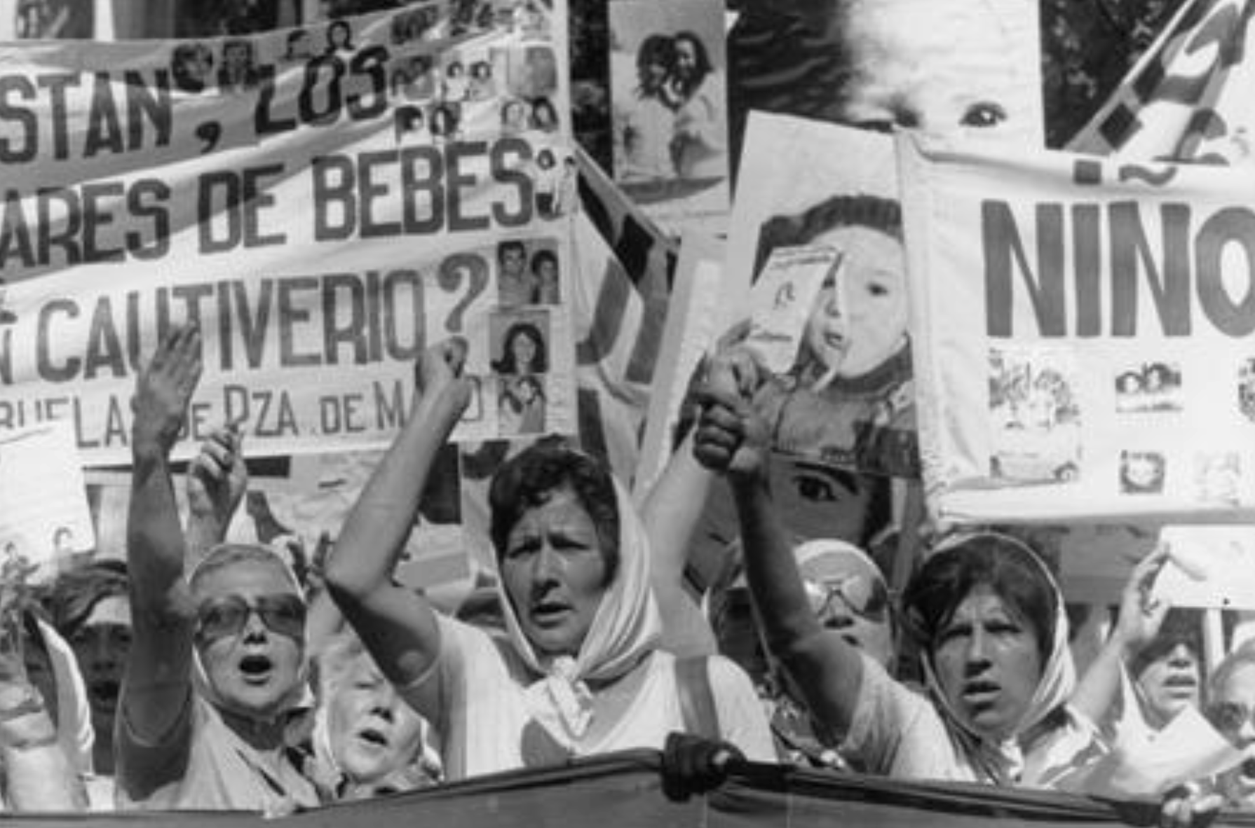 A group of women wearing white headscarves raise their fists and hold banners with photos of children and text during a protest.