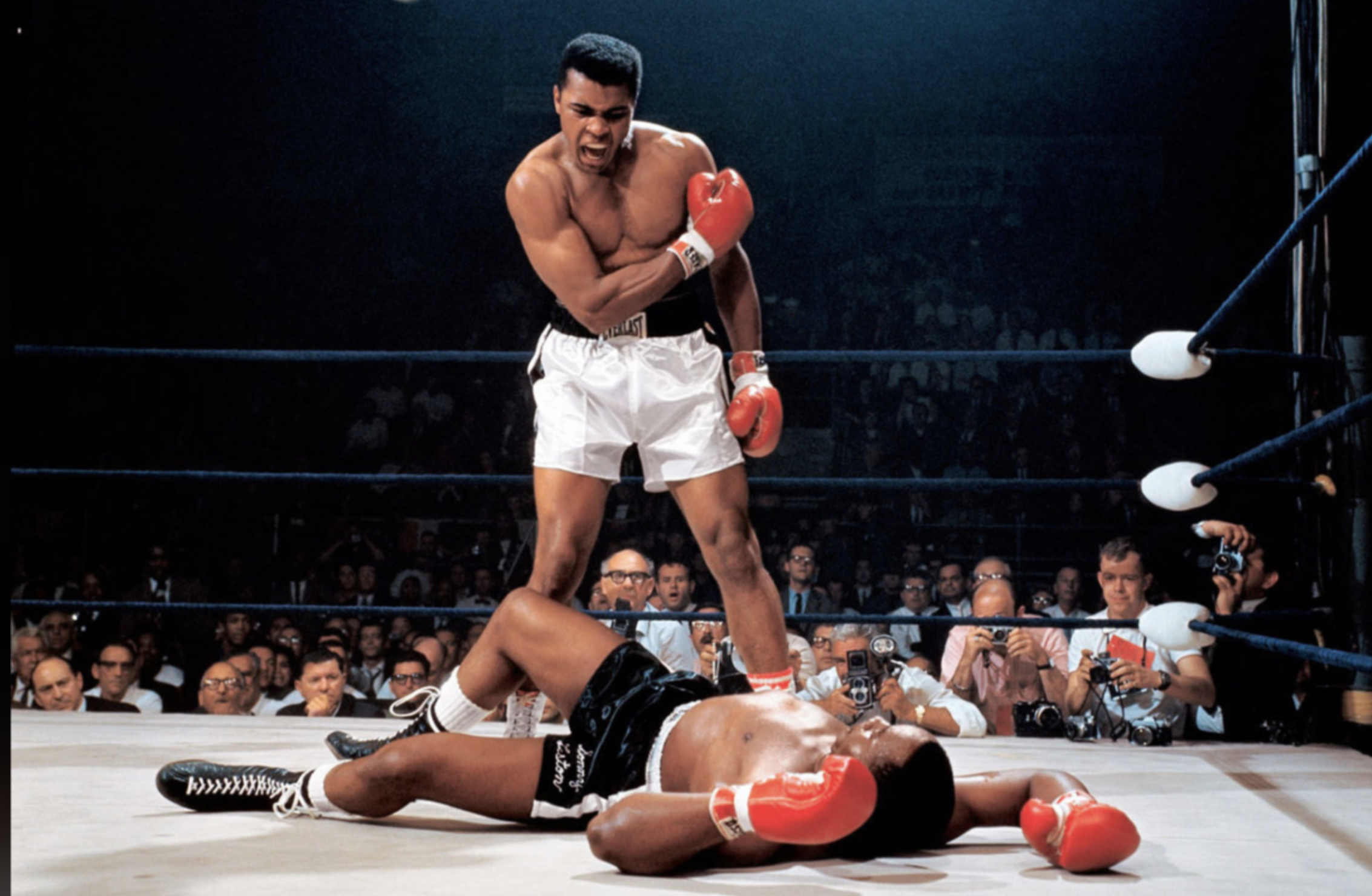 A boxer in white trunks and red gloves stands over his opponent, who is lying on the ring floor. The standing boxer is flexing his arm and yelling, surrounded by a crowd of spectators and photographers.