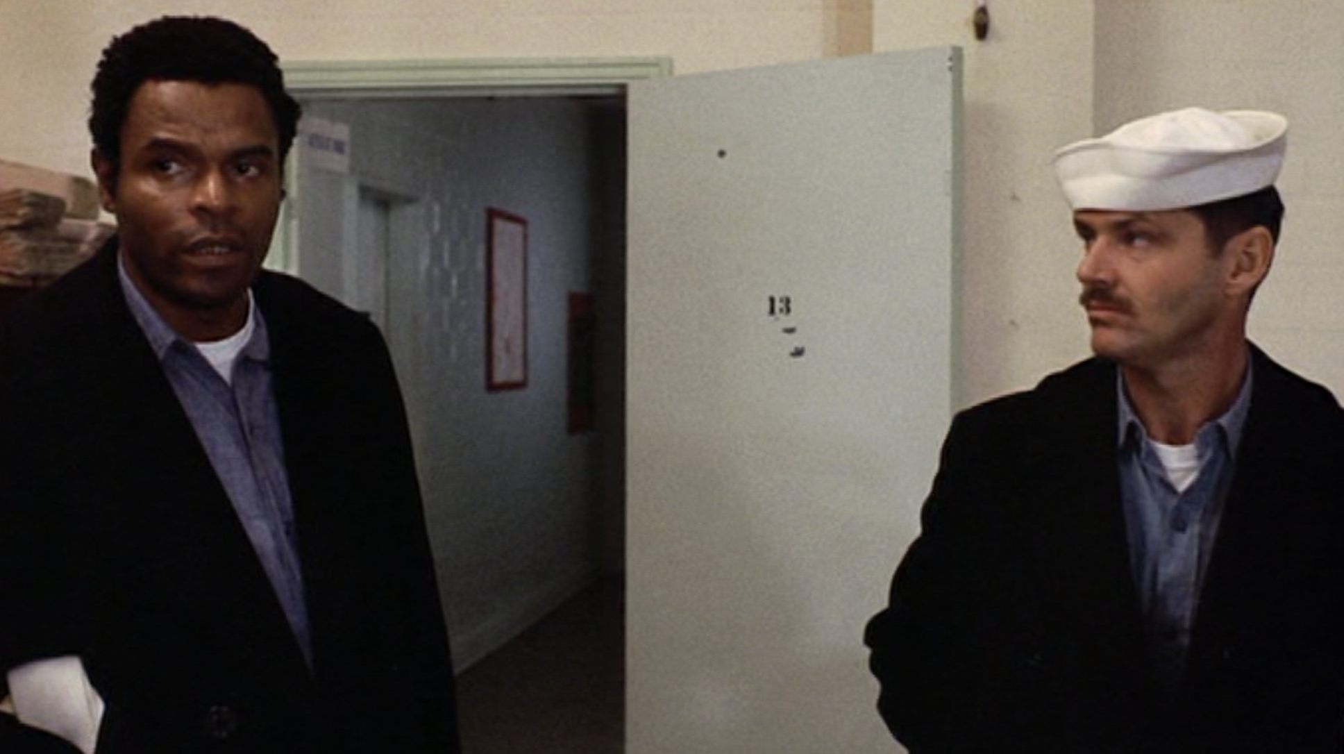 Two men stand in a hallway; one wears a dark jacket and blue shirt, the other wears a white sailor hat and uniform. An open white door with the number 13 is in the background. Both men look serious.