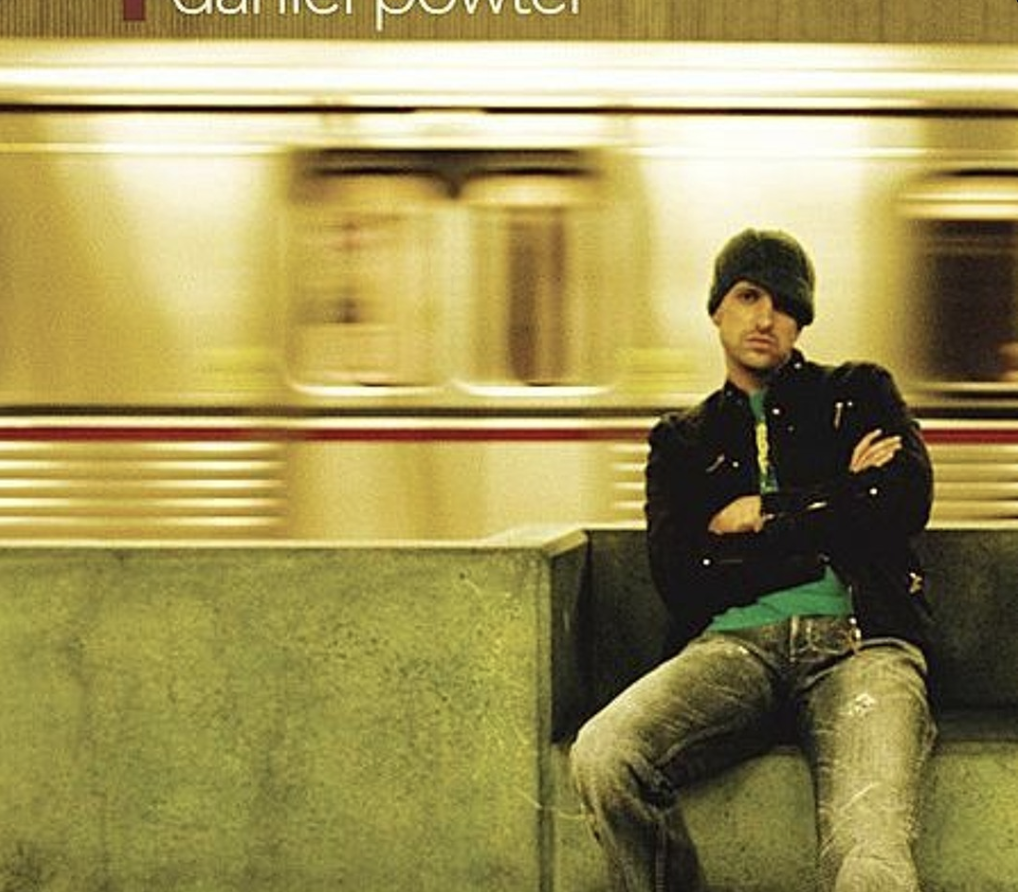 A man wearing a beanie and casual clothes sits on a bench with his arms crossed, while a subway train speeds by in the background. The scene has a motion blur effect.