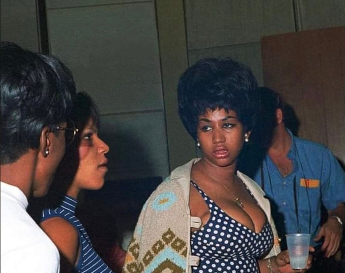 Three people stand indoors talking; a woman in the center with a stylish updo and polka-dot dress looks to her right, holding a cup, while two others engage in conversation.