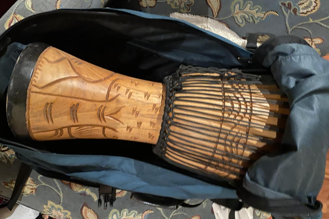 A wooden djembe drum with carved patterns is partially packed inside an open blue backpack, resting on a patterned fabric surface.