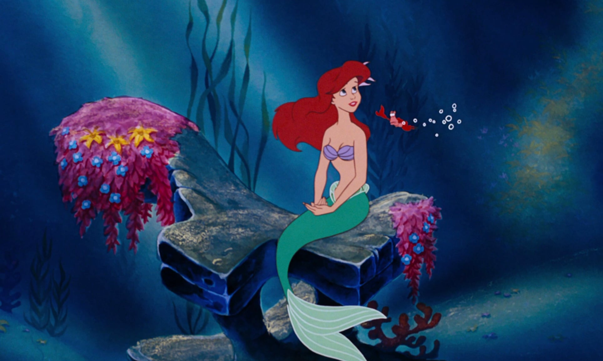 A red-haired mermaid in a purple seashell top and green tail sits on a rock underwater, surrounded by coral, with a small red crab blowing bubbles beside her.