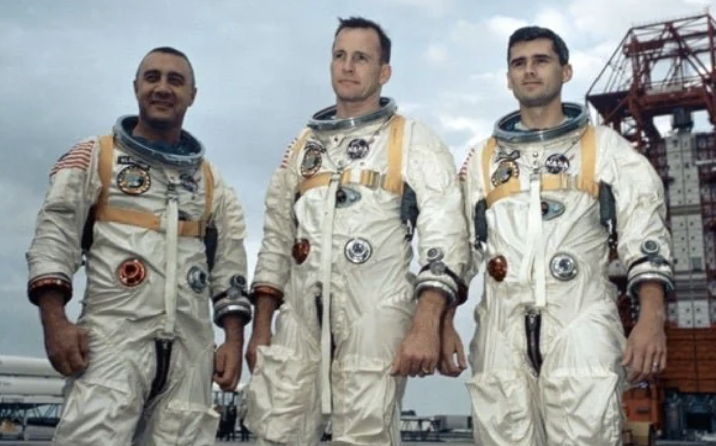 Three astronauts in white NASA spacesuits stand side by side outdoors near launch equipment, looking ahead. The sky is cloudy, and the astronauts appear ready for a mission.