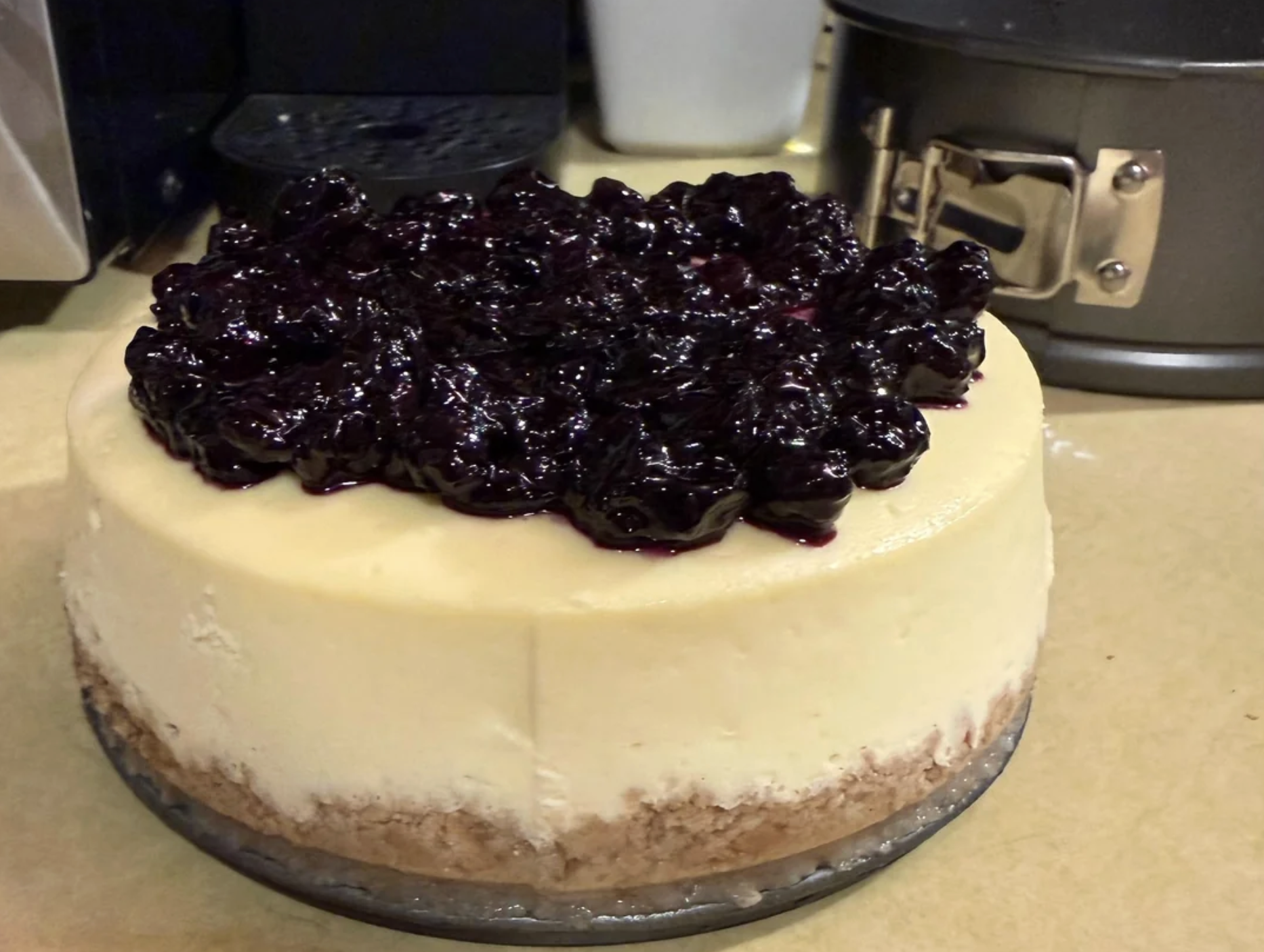 A homemade cheesecake with a thick graham cracker crust and creamy filling, topped with a generous layer of dark blueberry compote, sits on a kitchen counter near a springform pan.