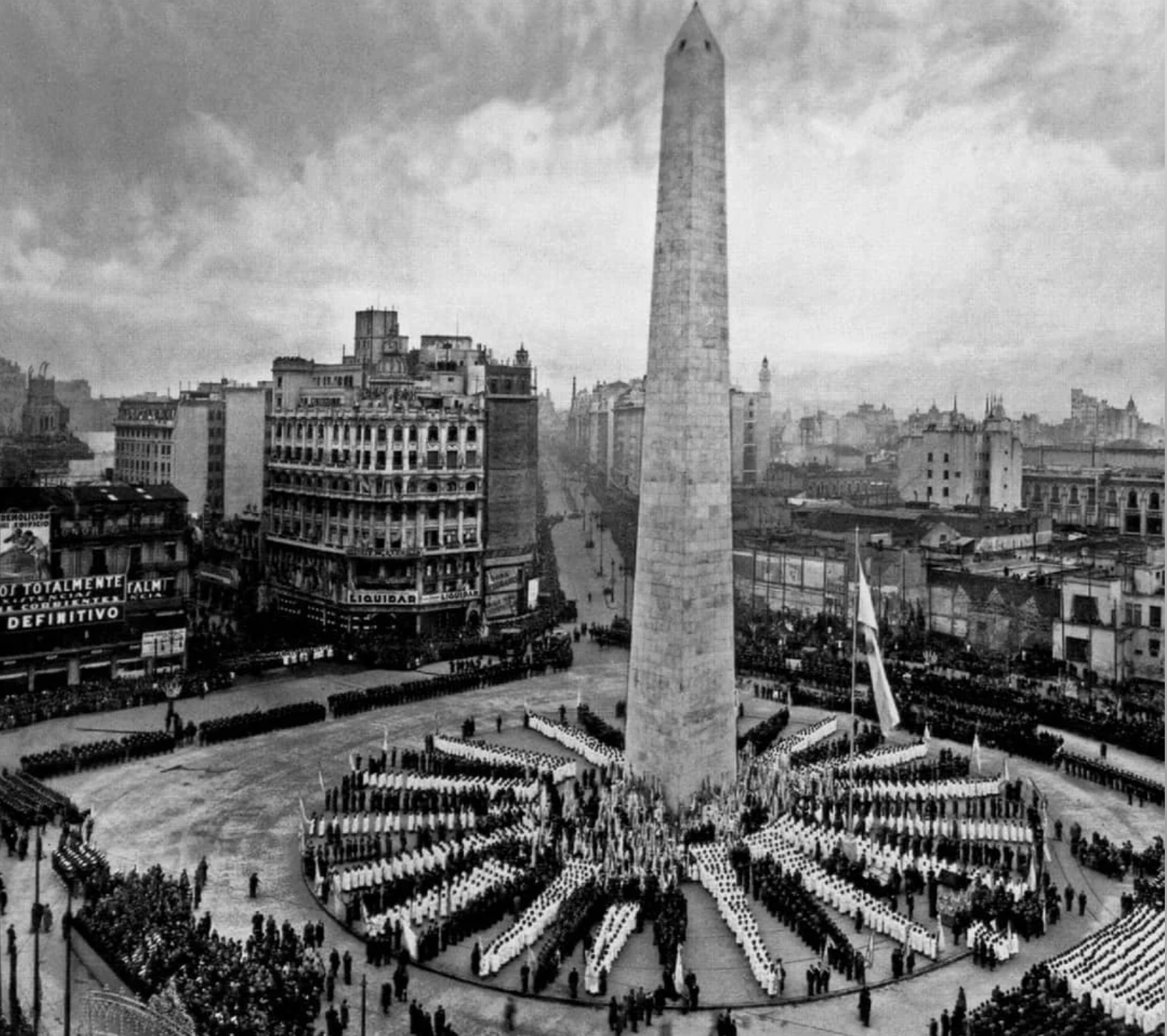 Black and white photo of a large crowd gathered around the Obelisk of Buenos Aires in a city square, with surrounding buildings and streets visible in the background.