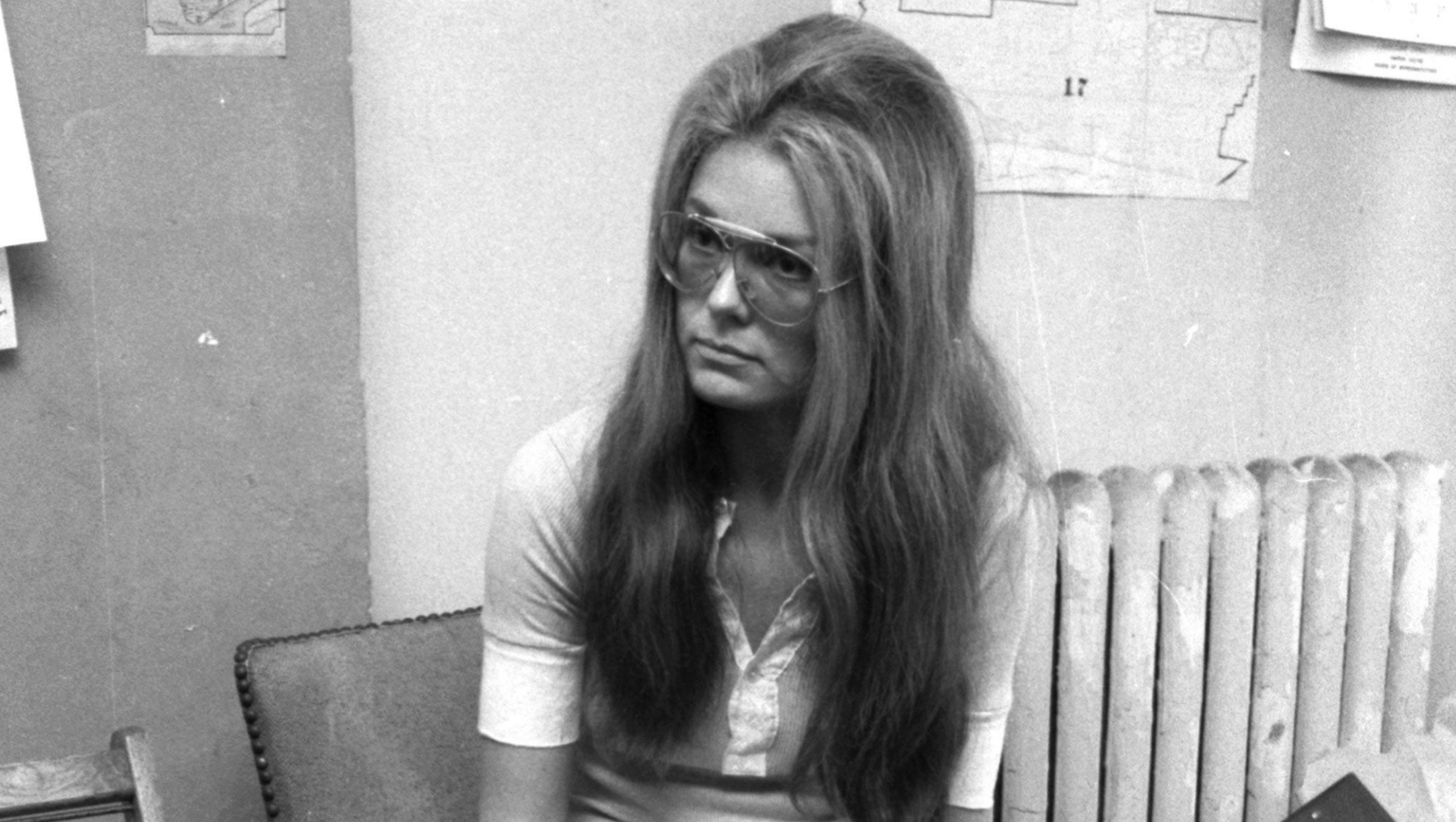 A woman with long hair and glasses sits indoors, wearing a light-colored shirt. A radiator, chair, and papers on the wall are visible in the background. The photo is in black and white.
