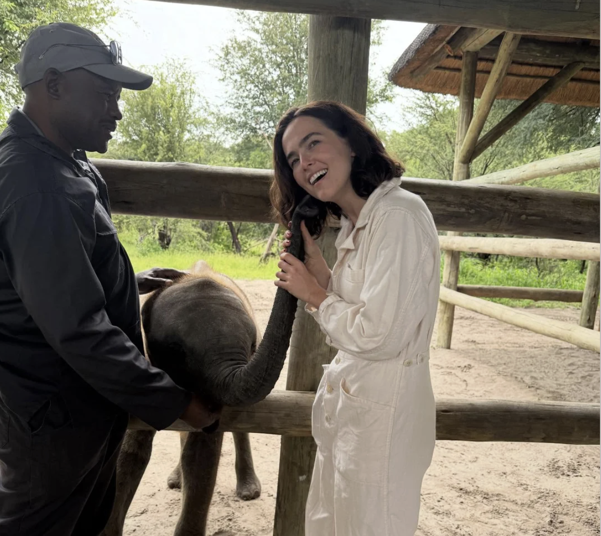 A woman in a white jumpsuit smiles while holding and nuzzling an elephant's trunk. A man in dark clothing stands nearby, guiding the elephant. They are in an outdoor, fenced area with greenery in the background.