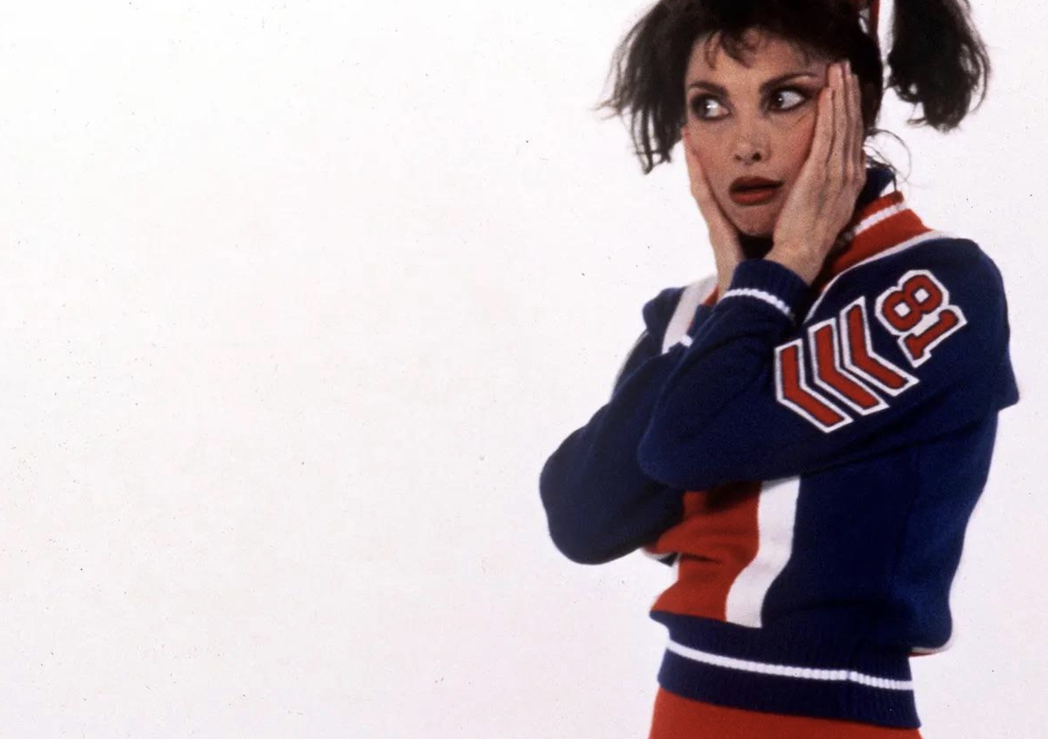 A woman in a blue, red, and white cheerleader uniform with the number 8 on the sleeve holds her face in surprise, with wide eyes and messy pigtails, standing against a plain white background.