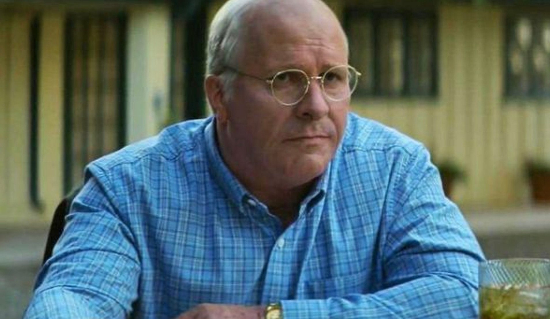 A middle-aged man with glasses and a bald head, wearing a blue plaid shirt, sits at a table outdoors with a drink in front of him, looking serious.