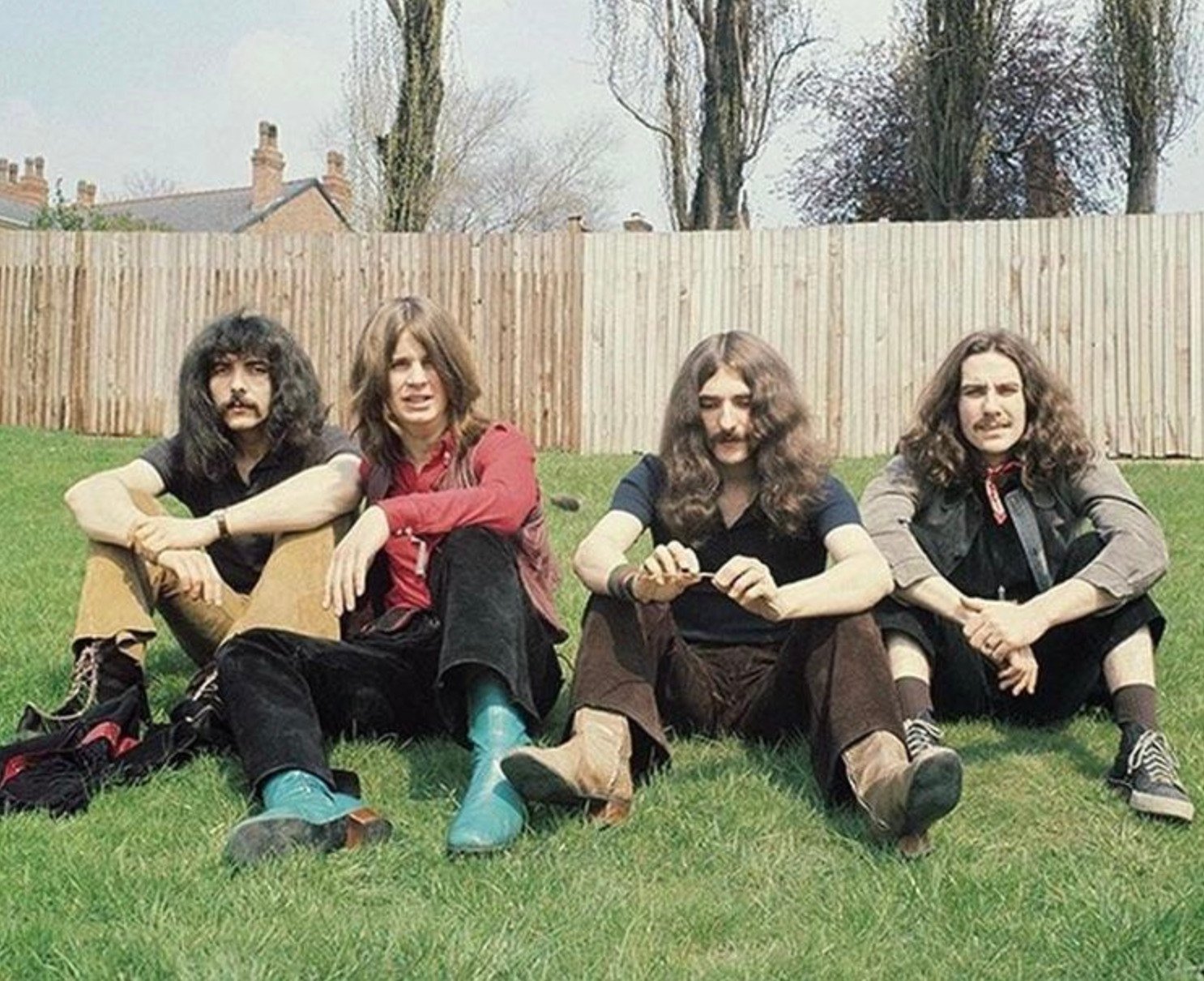 Four men with long hair sit on green grass in front of a wooden fence and trees. They wear vintage clothing, including boots and bell-bottoms, and look toward the camera in a relaxed pose.