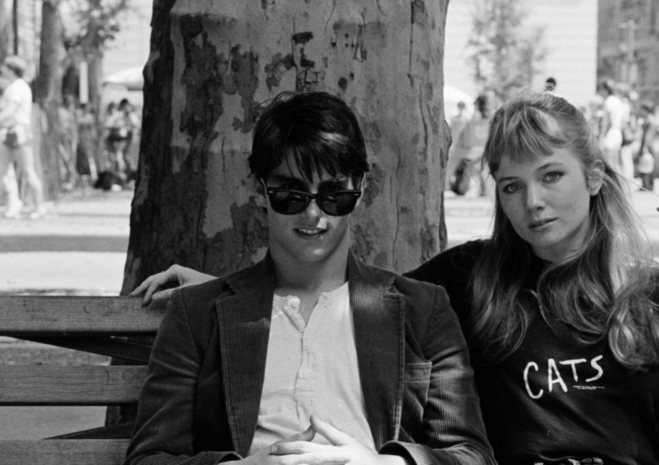 A young man in sunglasses and a jacket sits on a bench next to a woman with long hair wearing a "CATS" sweatshirt. They are in a park with a tree behind them and people in the background.