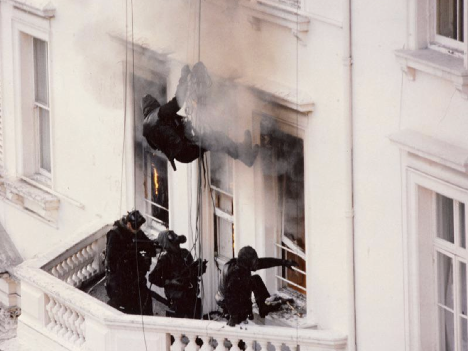 Armed officers in black uniforms abseil from the roof and enter a white building through windows as smoke billows out; more officers are positioned on a balcony below, appearing to prepare for entry.