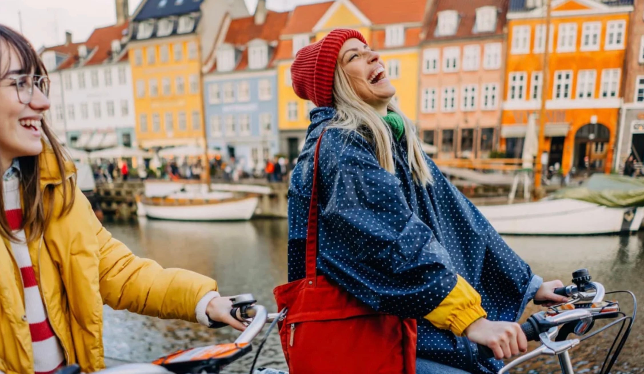 Two women laugh while riding bikes along a canal lined with colorful buildings. One wears a red beanie and navy poncho, the other a yellow jacket. Boats and people are visible in the lively background.